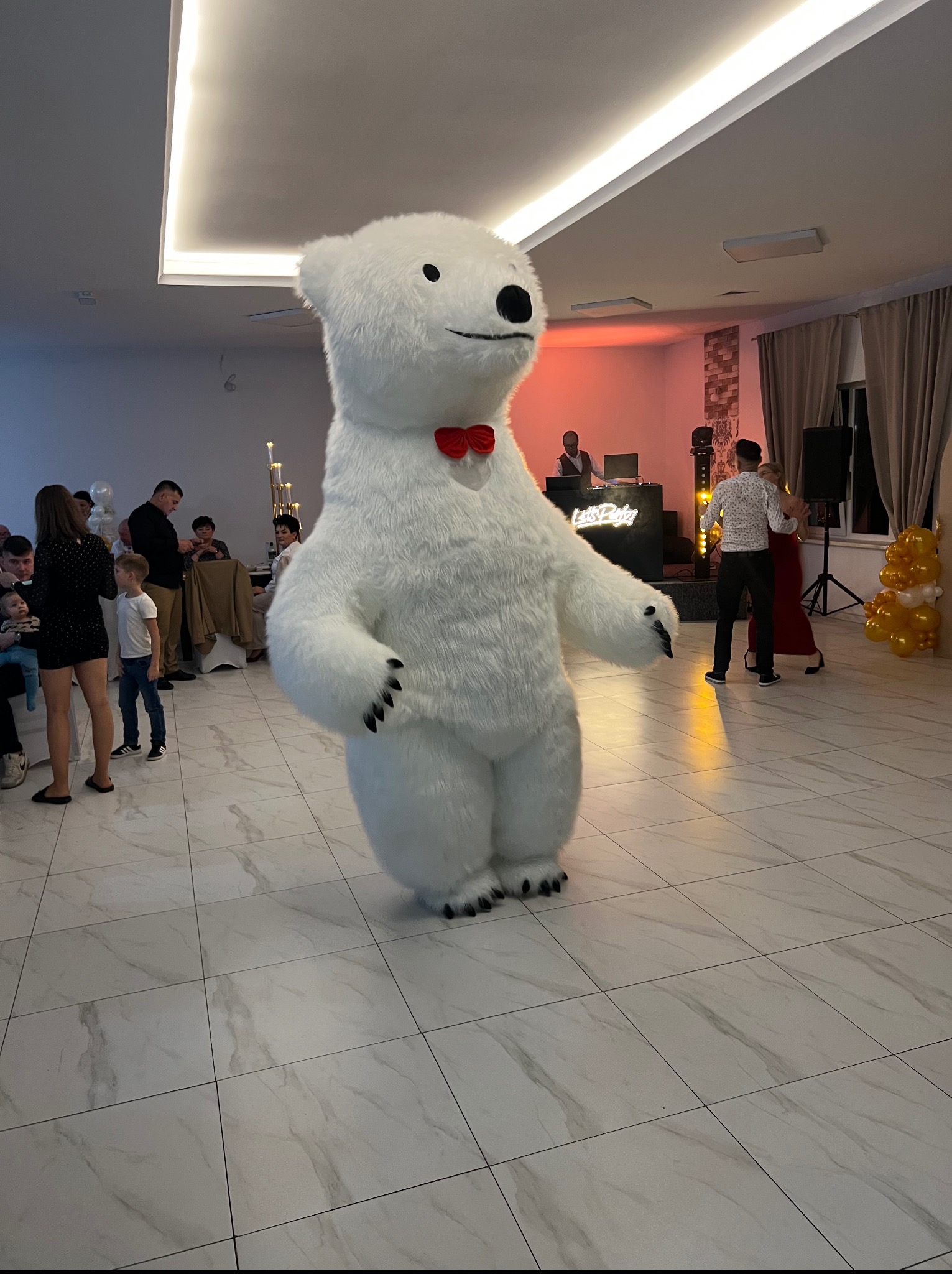 Duży, pluszowy biały niedźwiedź z czerwoną muszką, stoi na parkiecie w sali balowej podczas imprezy. W tle DJ i tańcząca para.