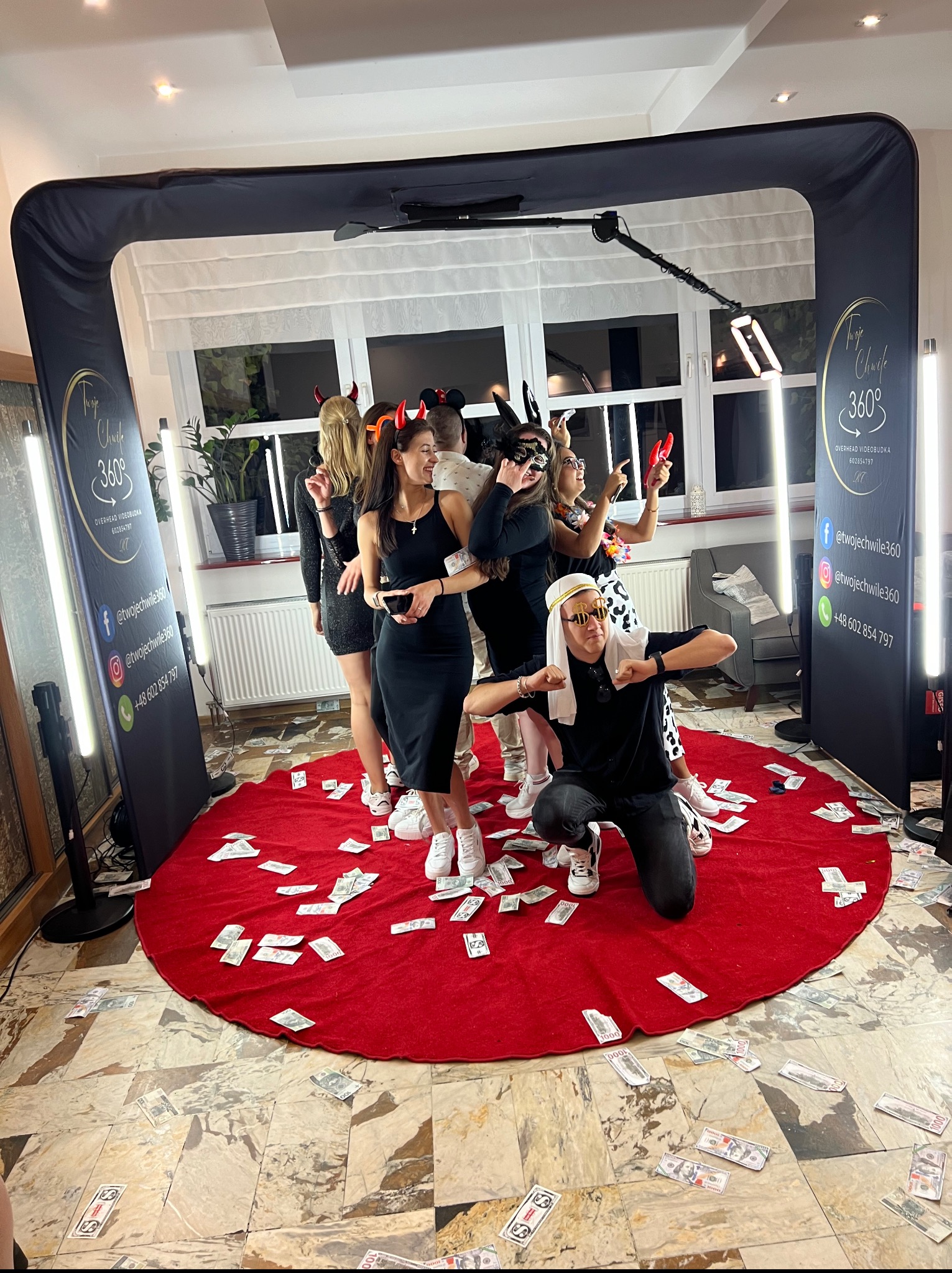 Wesoła grupa w przebraniach pozuje w videobudce 360° na czerwonym dywanie zasypanym rekwizytami imitującymi pieniądze. Na ściankach budki widoczne logo i dane kontaktowe.