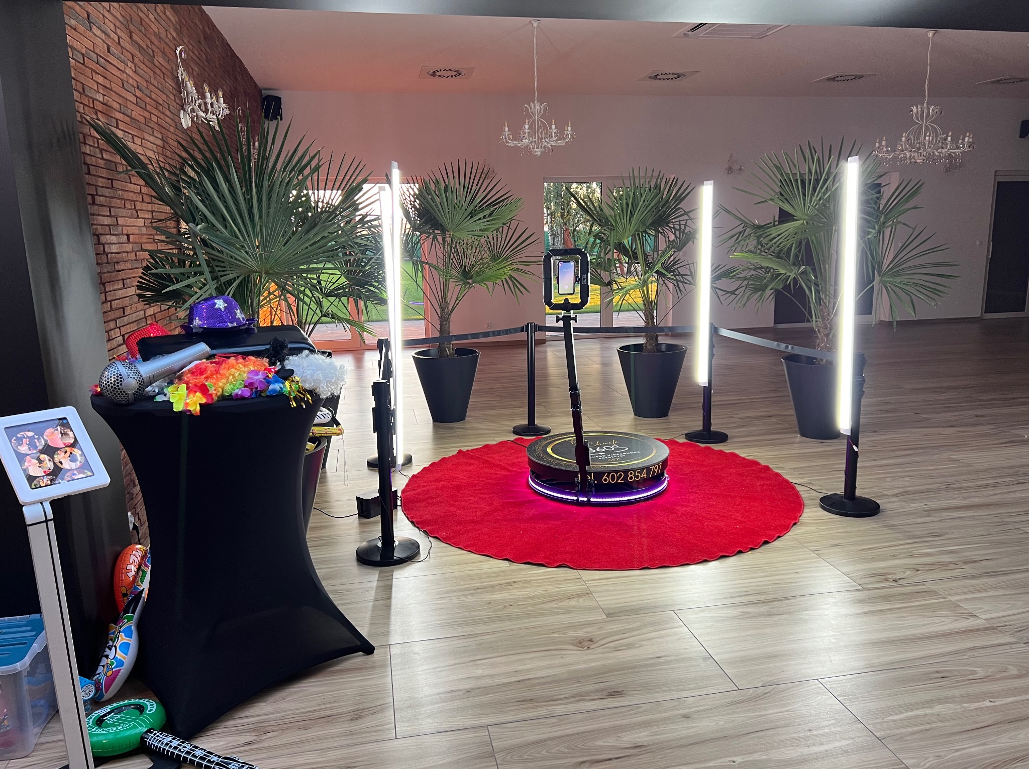 Videobudka 360 na czerwonym dywanie, otoczona palmami i lampami. Dodatkowo stolik z akcesoriami do zdjęć. Przestronne wnętrze z parkietem i żyrandolami.