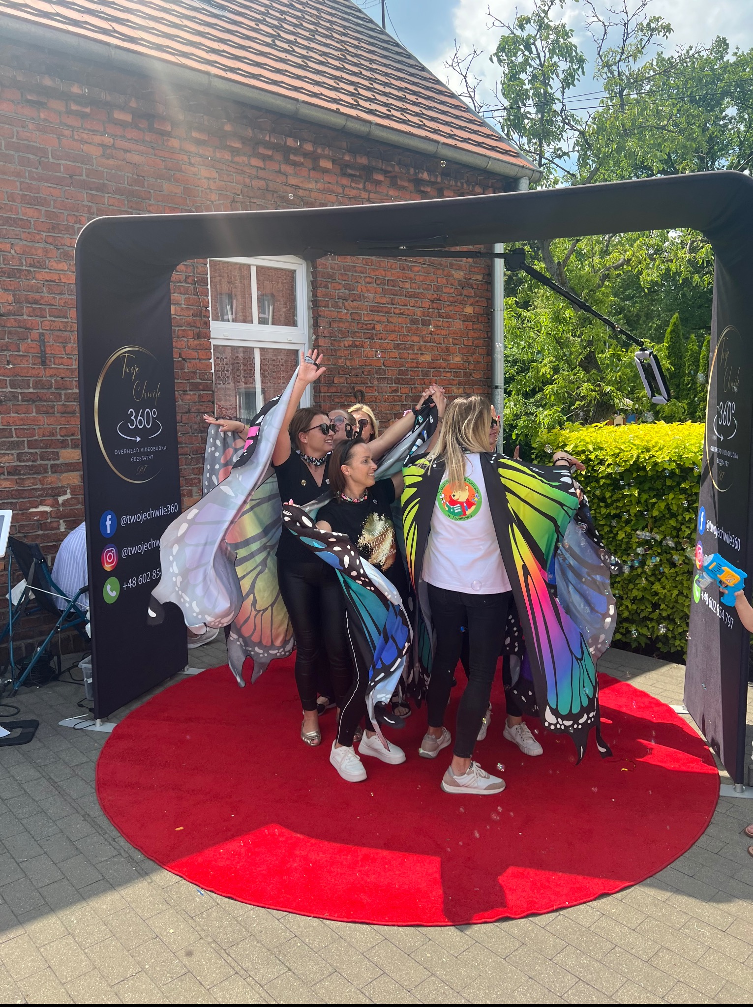 Videobudka 360 w Słupcy: grupa kobiet w kolorowych skrzydłach pozuje na czerwonym dywanie. W tle budynek z cegły i zieleń. Zabawa i uśmiech na evencie.
