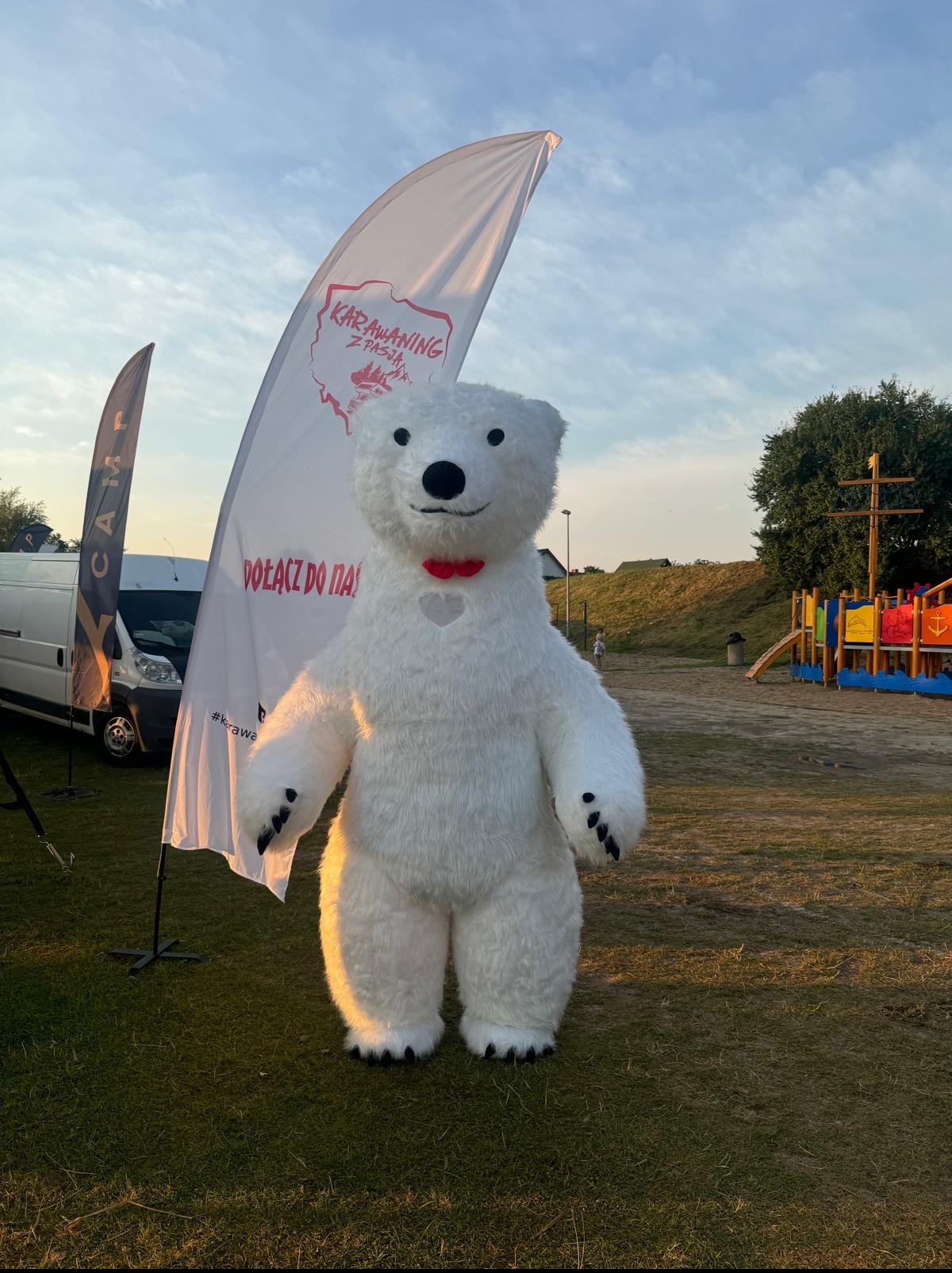 Duży, biały miś w kostiumie na tle flagi 'Karawaning z pasją' i placu zabaw. Maskotka z czerwoną muchą.