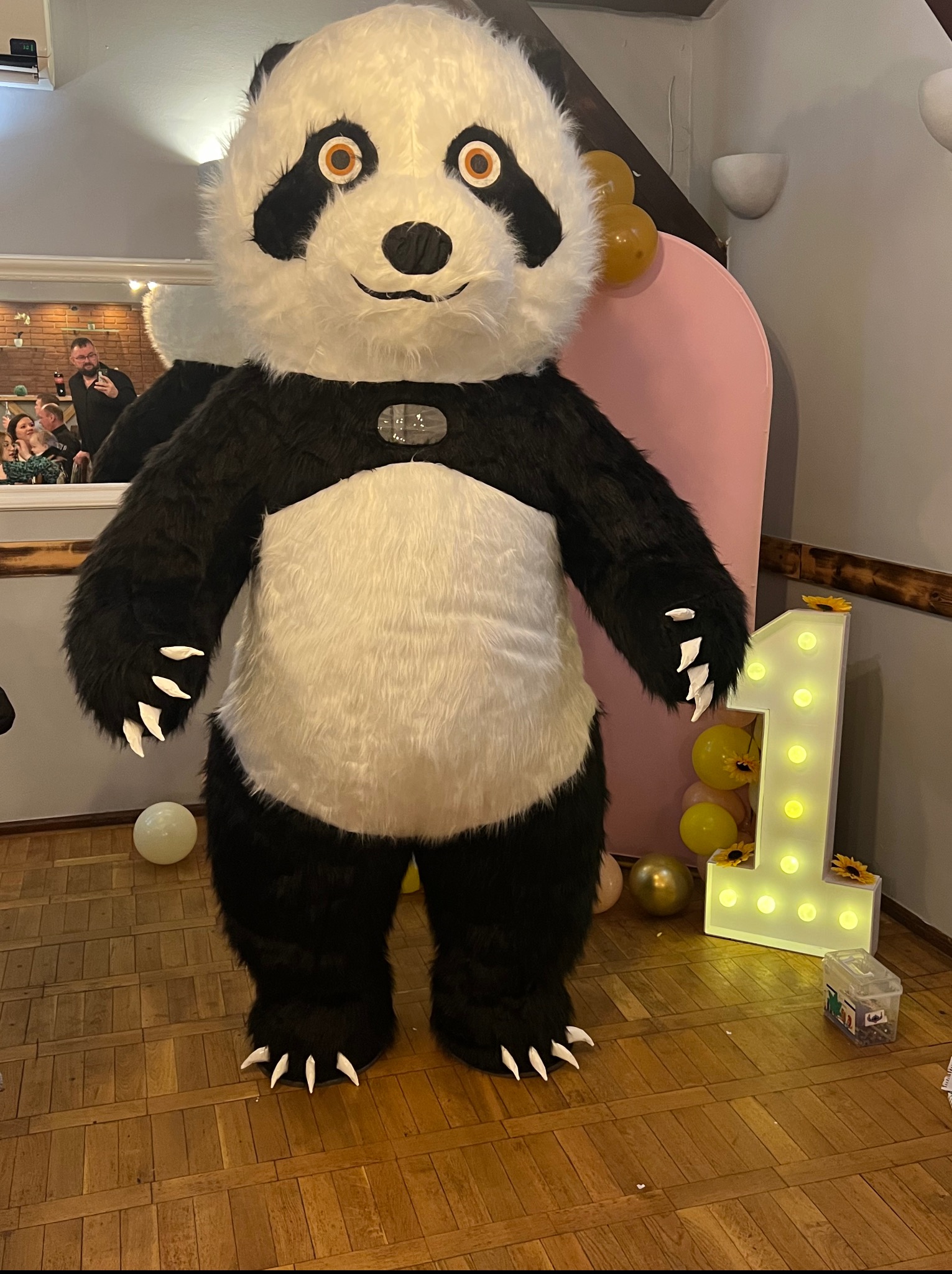 Panda, maskotka na urodzinach dziecka, stoi obok podświetlanej cyfry '1' i balonów. W tle lustro odbija bawiących się gości.