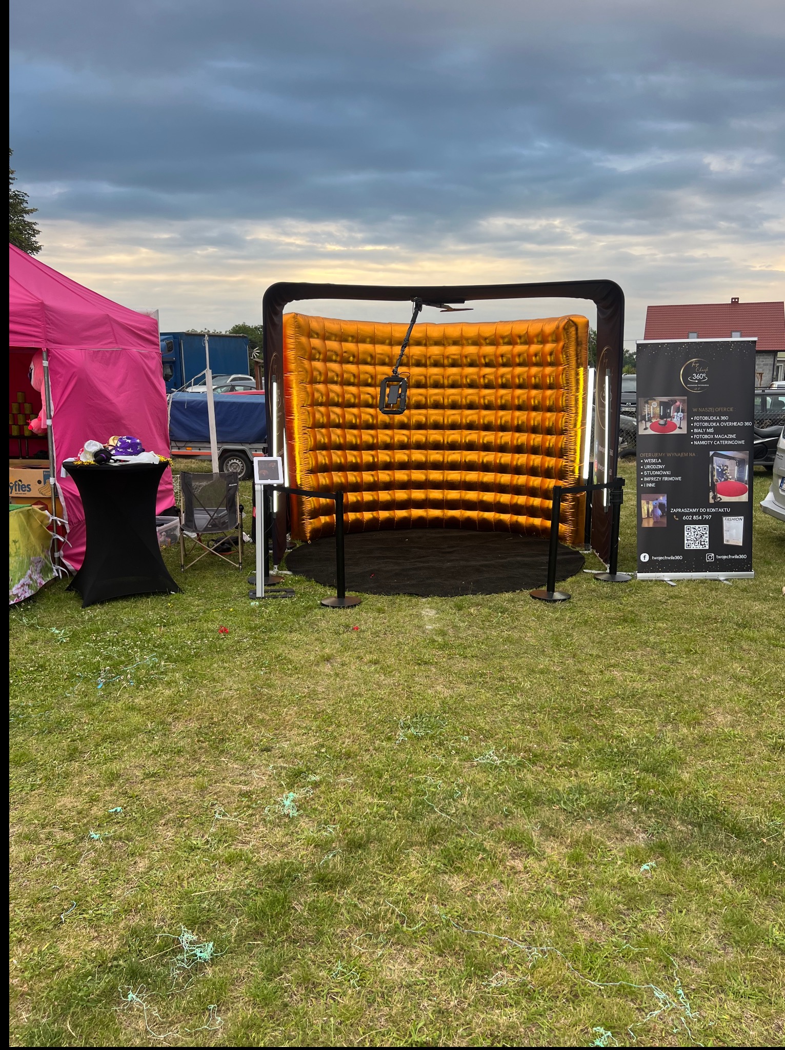 Złota fotobudka 360 na trawie, obok różowy namiot i baner reklamowy. Dekoracje rozsypane na ziemi. Pochmurne niebo w tle, widok z zewnątrz.