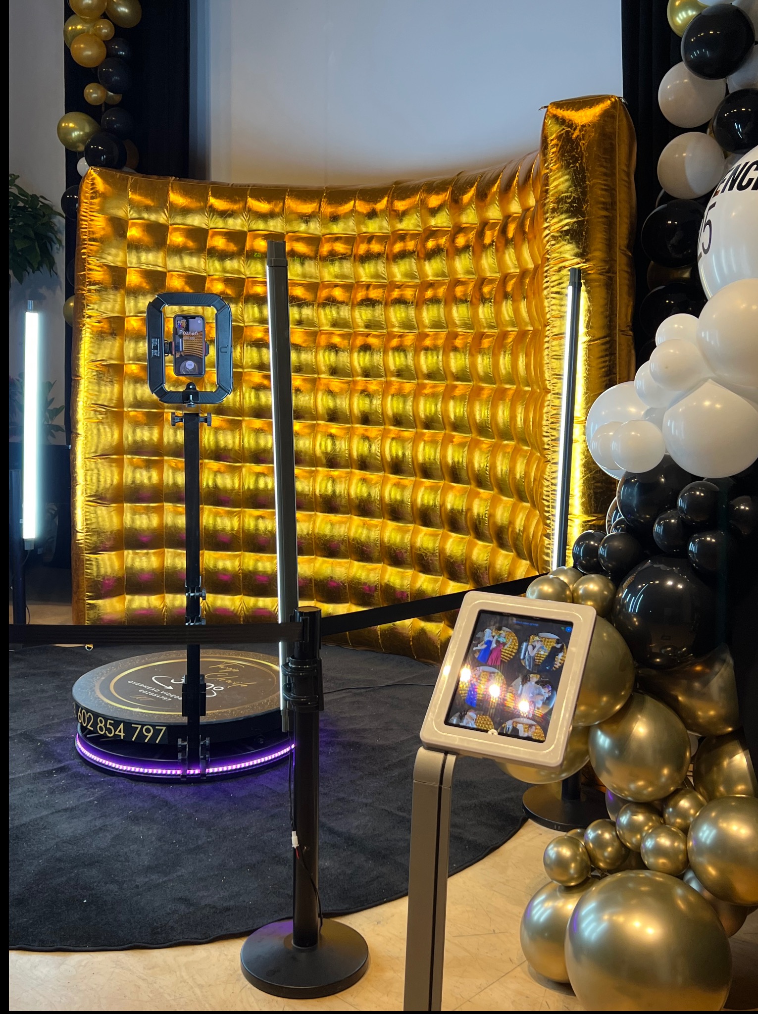 Złota ścianka, platforma obrotowa 360 stopni i stacja sharingowa z podglądem nagrań na tle czarno-złotych balonów. Nowoczesna fotobudka na imprezę.