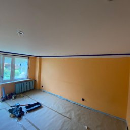 Dawid Szklanowski - Pokój w trakcie malowania na pomarańczowo, zabezpieczone okna folią, listwy sufitowe z taśmą, podłoga zakryta folią malarską, widoczne narzędzia malarskie.