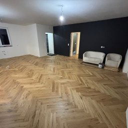 Dawid Szklanowski - Ukończone układanie parkietu w jodełkę w jasnym odcieniu drewna, czarna ściana, białe ściany, dwa fotele i elementy wykończeniowe w pomieszczeniu.