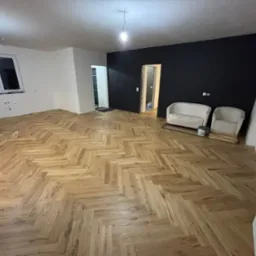 Ukończone układanie parkietu w jodełkę w jasnym odcieniu drewna, czarna ściana, białe ściany, dwa fotele i elementy wykończeniowe w pomieszczeniu.