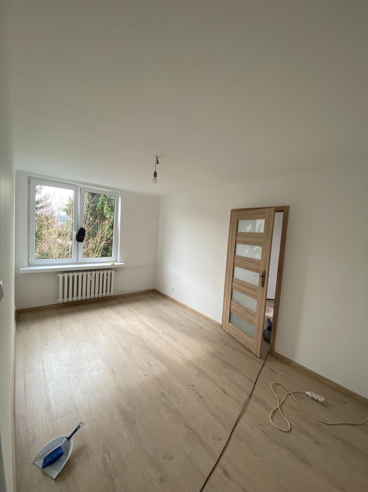 Jasne wnętrze po remoncie: świeżo pomalowane ściany, drewniana podłoga, okno z widokiem na zieleń. Otwarta, drewniana  futryna drzwi z przeszkleniami.