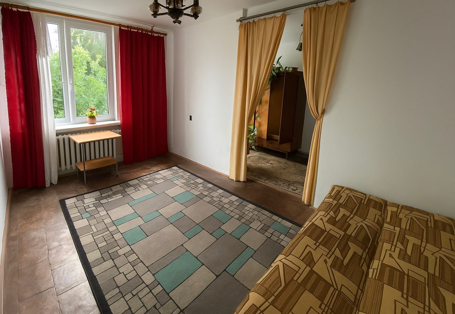 Ustawny pokój z dywanem w geometryczne wzory, czerwonymi zasłonami przy oknie i beżowymi w przejściu do kolejnego pomieszczenia. Widoczny stolik i fragment sofy.