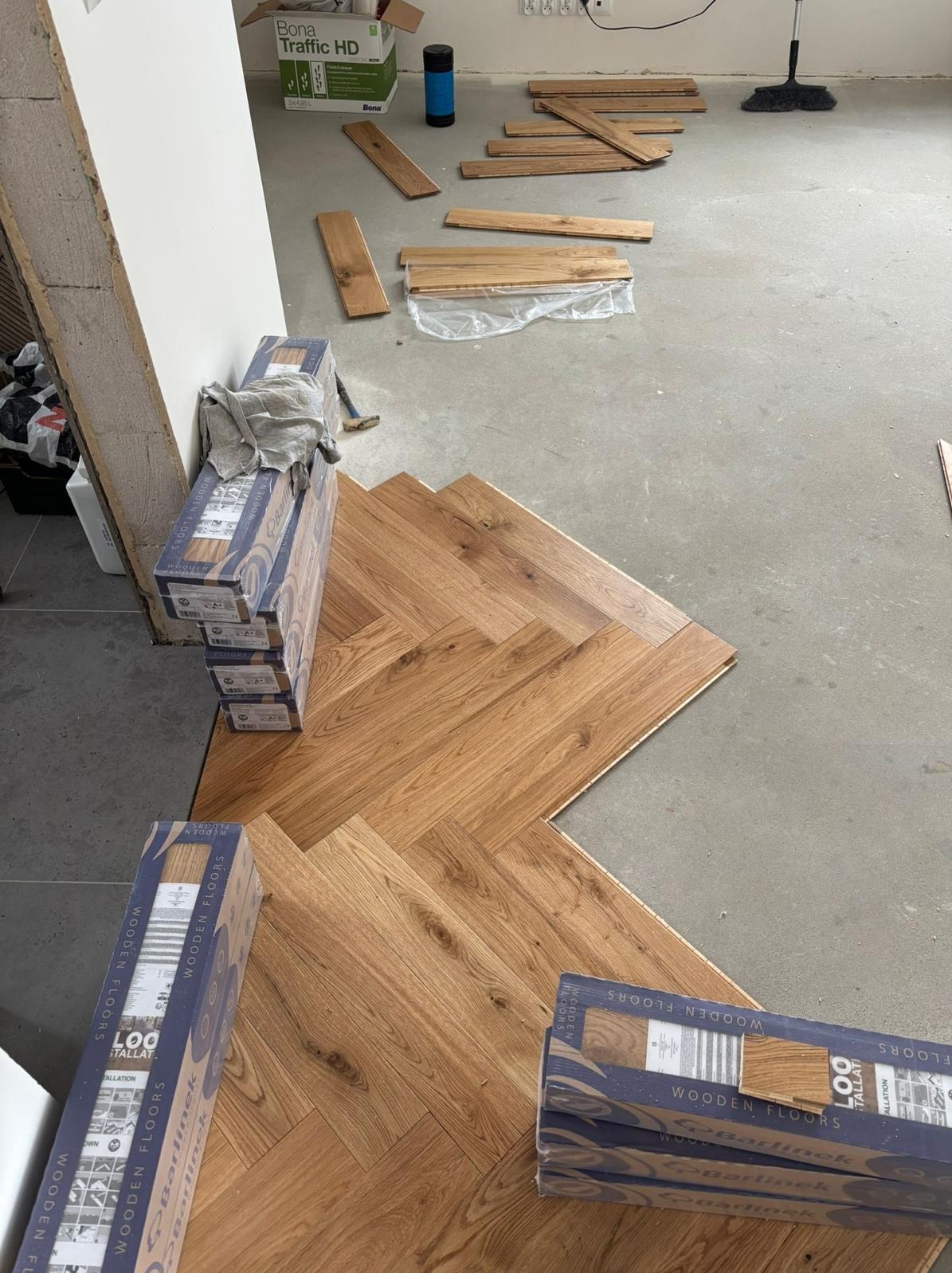 Układanie parkietu w jodełkę: częściowo ułożona podłoga z drewnianych paneli Barlinek, paczki paneli obok, przygotowane deski i narzędzia w tle.