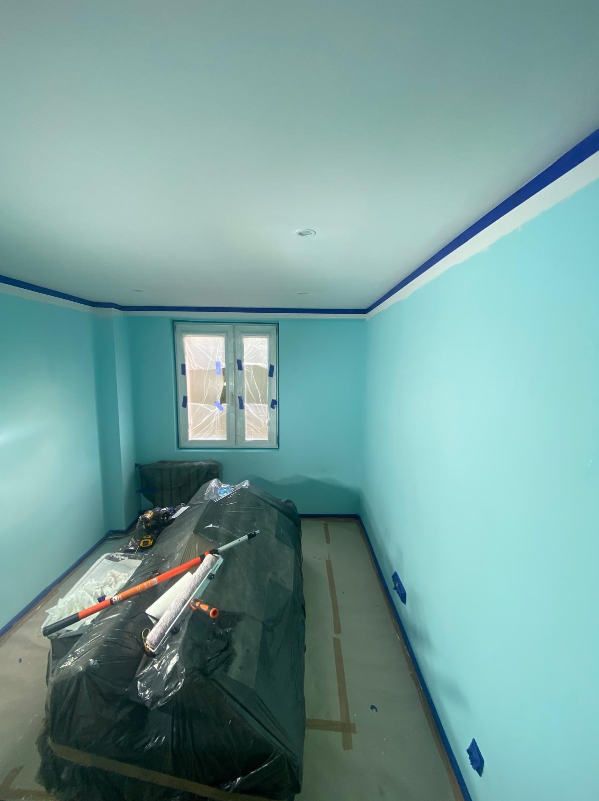 Pomieszczenie pomalowane na błękitno, zabezpieczone taśmą malarską przy suficie i podłodze. Okno zakryte folią. Sprzęt malarski przykryty folią ochronną.