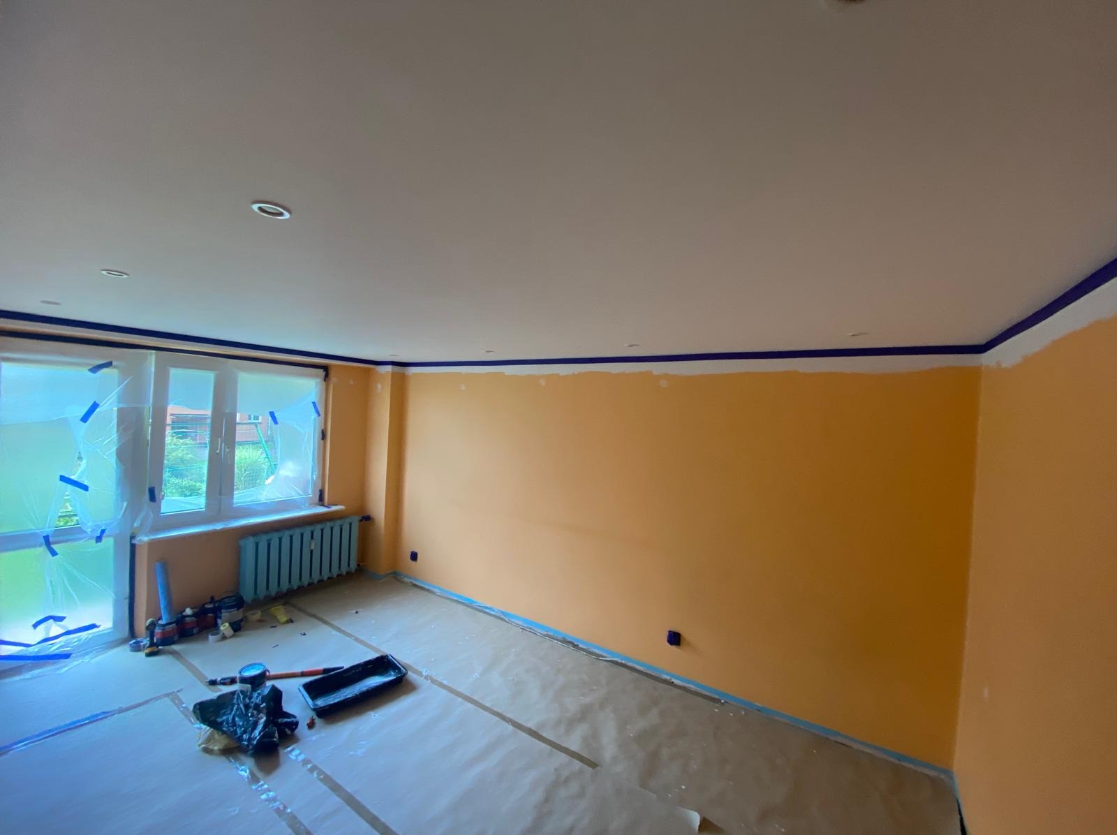 Pokój w trakcie malowania na pomarańczowo, zabezpieczone okna folią, listwy sufitowe z taśmą, podłoga zakryta folią malarską, widoczne narzędzia malarskie.