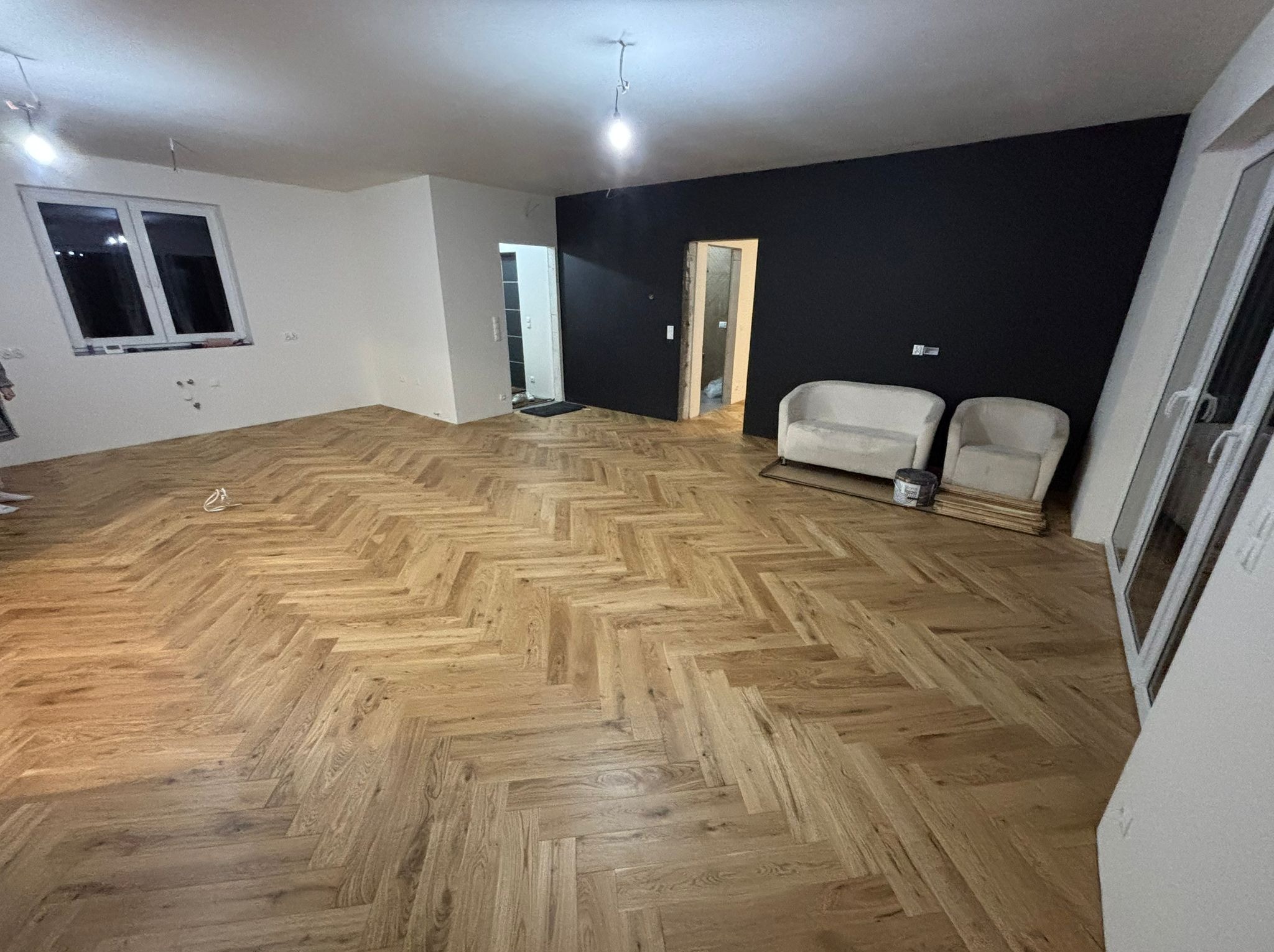 Ukończone układanie parkietu w jodełkę w jasnym odcieniu drewna, czarna ściana, białe ściany, dwa fotele i elementy wykończeniowe w pomieszczeniu.