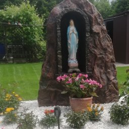 Kompleksowe Usługi Rzeszów - Grotka z figurą Matki Boskiej w niebiesko-białych szatach, na tle zielonego trawnika. Przed nią rabata z białych kamieni, różowymi hortensjami w donicy, żółtymi kwiatami i solarną lampką ogrodową.