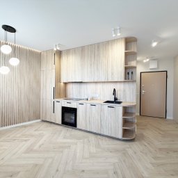 Fajne Remonty - Jasna, nowoczesna kuchnia z drewnianymi frontami i parkietem w jodełkę. Minimalistyczne oświetlenie i zabudowa meblowa tworzą spójną, przestronną aranżację wnętrza.
