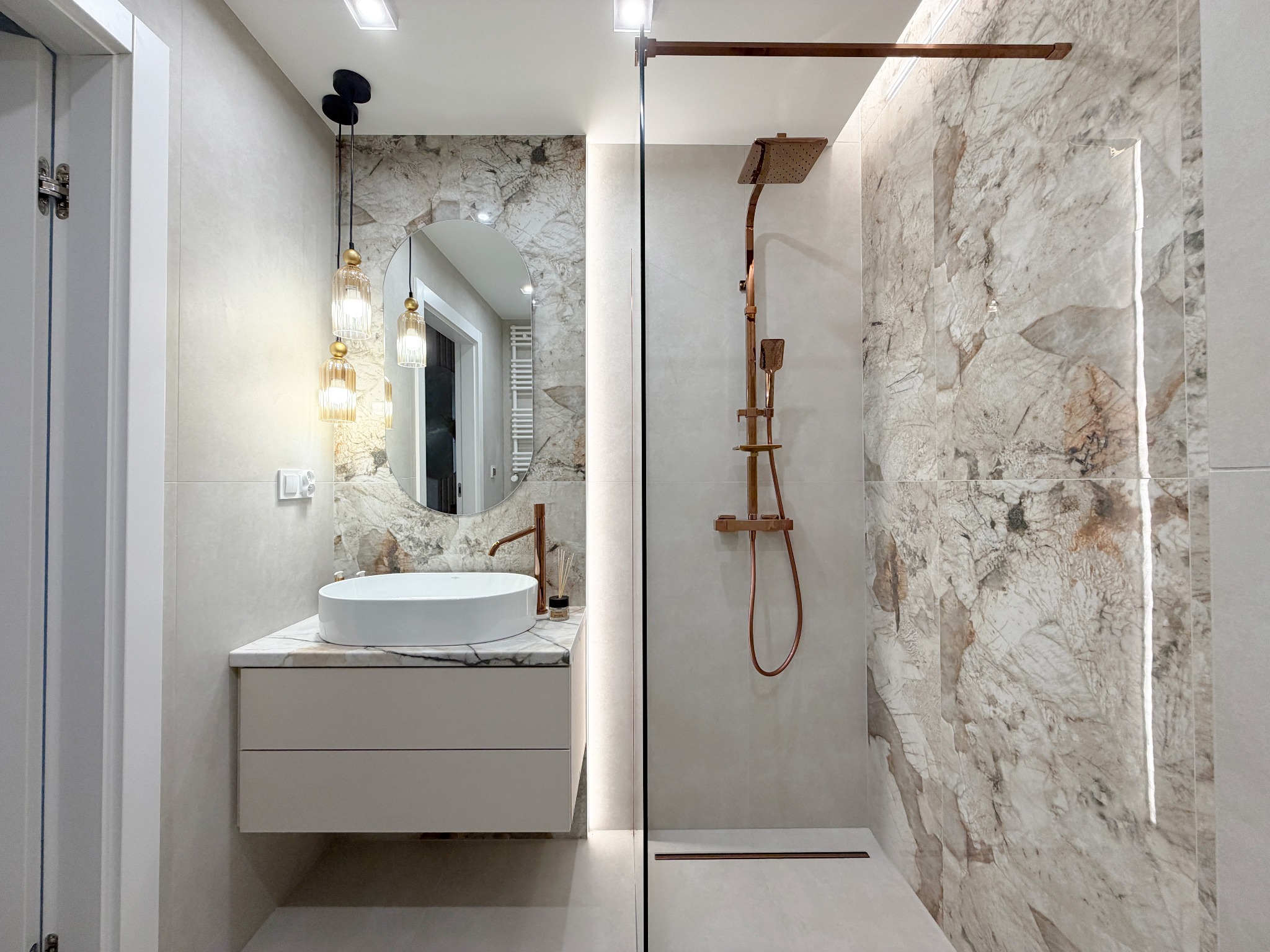 Elegancka łazienka po remoncie: kabina walk-in z miedzianą armaturą, umywalka nablatowa na szafce z marmurowym blatem, okrągłe lustro i wiszące lampy.