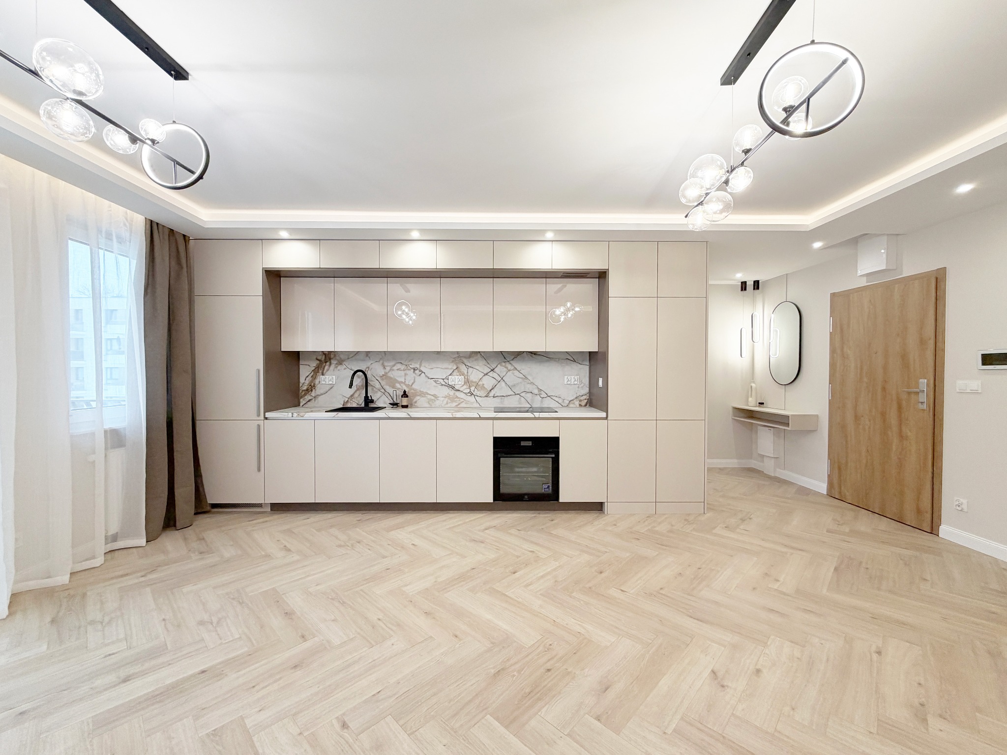 Jasne, przestronne wnętrze z parkietem w jodełkę, beżową zabudową kuchenną z marmurowym blatem i nowoczesnym oświetleniem. Widok na przedpokój z lustrem i konsolą.