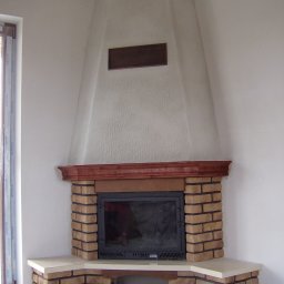 Jarosław Boder - Kanciasty kominek z cegły klinkierowej, drewnianym blatem i trapezowym wykończeniem ściany, w rogu pomieszczenia z oknem. Widok na surowy sufit.