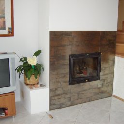 Jarosław Boder - Obudowa kominka z ciemnych, metalizowanych płytek, z widocznym paleniskiem. Obok drewniane schody i telewizor z kwiatem. Jasna, kwadratowa podłoga z płytek.