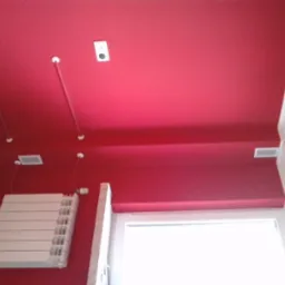 Czerwony sufit i ściany z białym grzejnikiem oraz rurami w pomieszczeniu po malowaniu. Widoczne kratki wentylacyjne i okno z roletą.