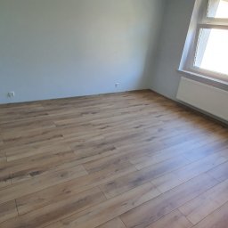 Jarosław Boder - Pokój po remoncie: panele podłogowe w odcieniach brązu, świeżo pomalowane szare ściany, biały grzejnik pod oknem. Widoczna taśma malarska na futrynie.