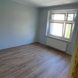 Jasne panele podłogowe w pokoju z oknem, świeżo po remoncie. Ściany pomalowane na szaro, widoczny biały grzejnik i sufit z dekoracyjnym oświetleniem.