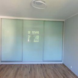 Jarosław Boder - Wnętrze pokoju w Gliwicach po remoncie: drewniana podłoga, szare ściany i zabudowa z przesuwnymi, mlecznymi drzwiami. Drzwi wejściowe zabezpieczone folią.