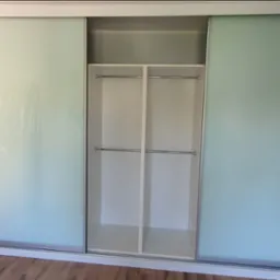 Biała szafa wnękowa z przesuwnymi drzwiami z mlecznego szkła, widoczne p&oacute;łki i drążki na ubrania. Podłoga z paneli.