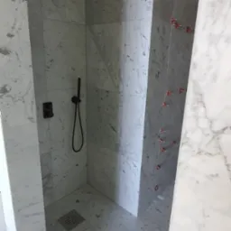 Wykończona kabina prysznicowa z marmurowymi płytkami, czarną armaturą i odpływem liniowym. Na ścianie widoczne krzyżyki dystansowe do płytek.