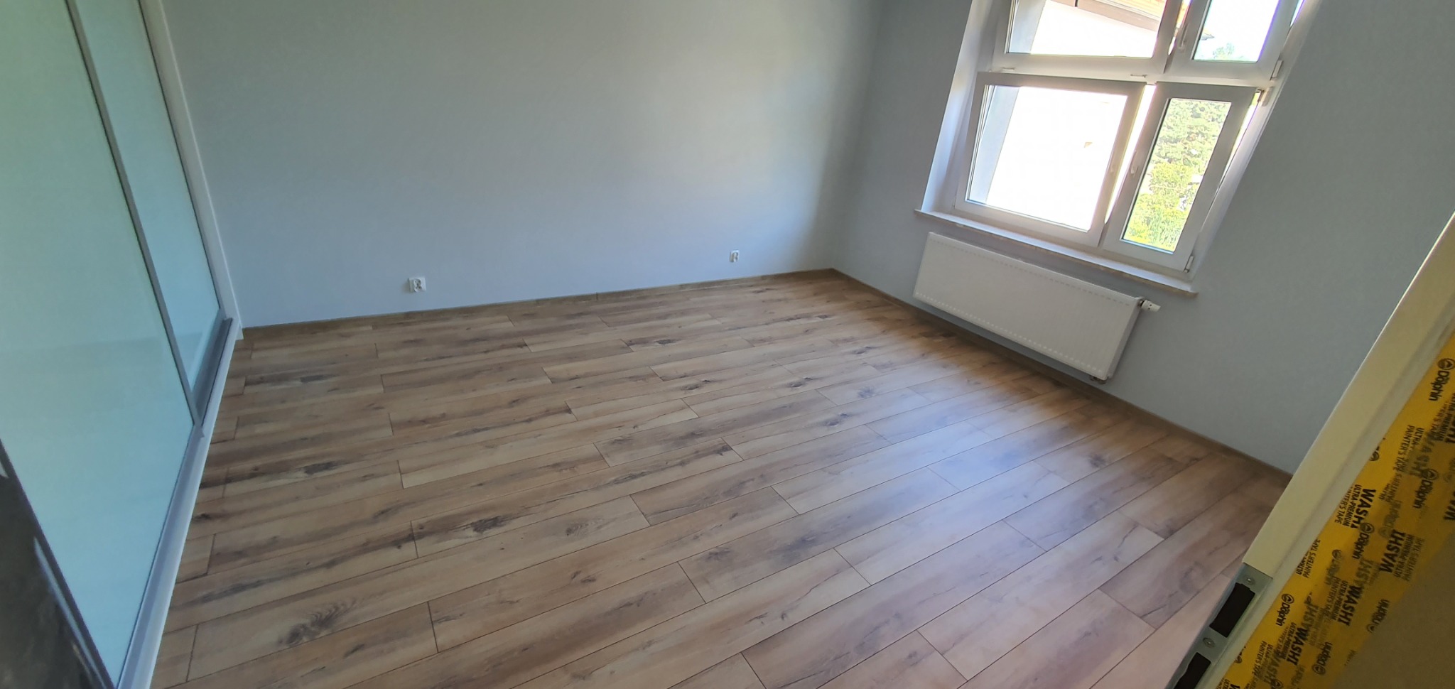 Pokój po remoncie: panele podłogowe w odcieniach brązu, świeżo pomalowane szare ściany, biały grzejnik pod oknem. Widoczna taśma malarska na futrynie.