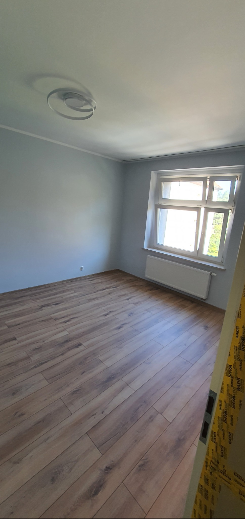 Jasne panele podłogowe w pokoju z oknem, świeżo po remoncie. Ściany pomalowane na szaro, widoczny biały grzejnik i sufit z dekoracyjnym oświetleniem.