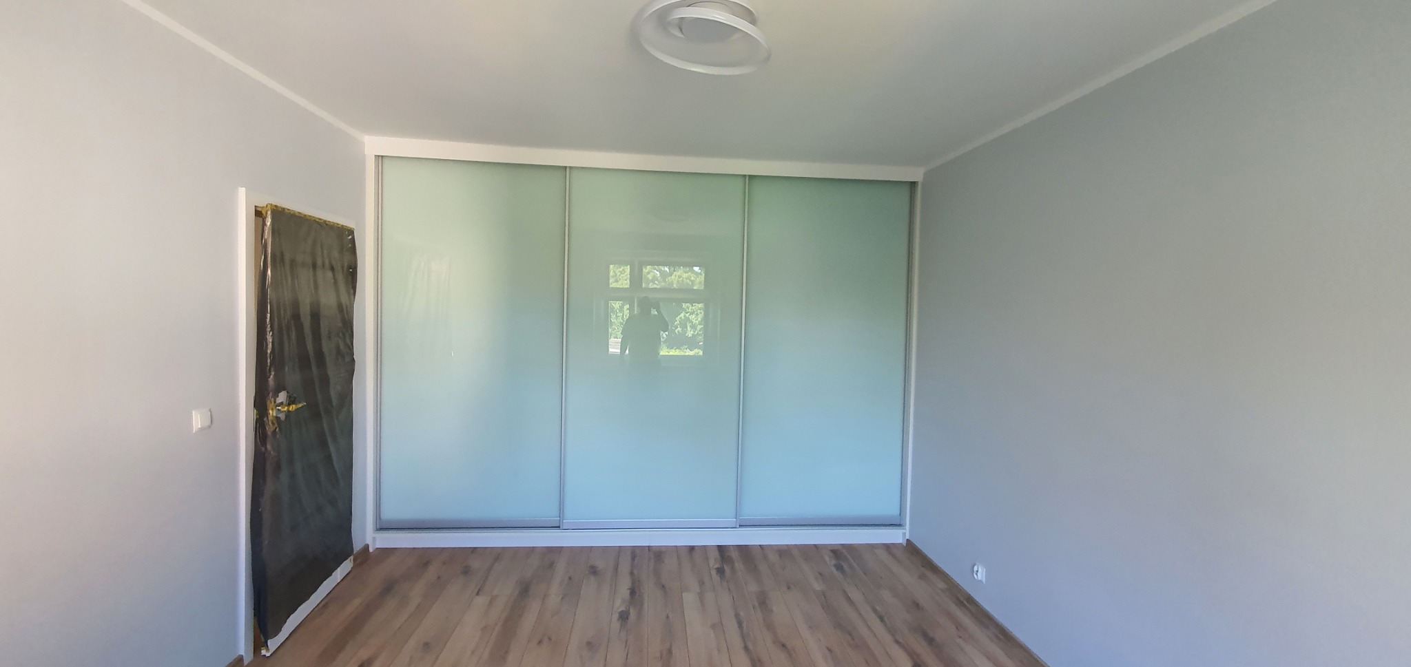 Wnętrze pokoju w Gliwicach po remoncie: drewniana podłoga, szare ściany i zabudowa z przesuwnymi, mlecznymi drzwiami. Drzwi wejściowe zabezpieczone folią.