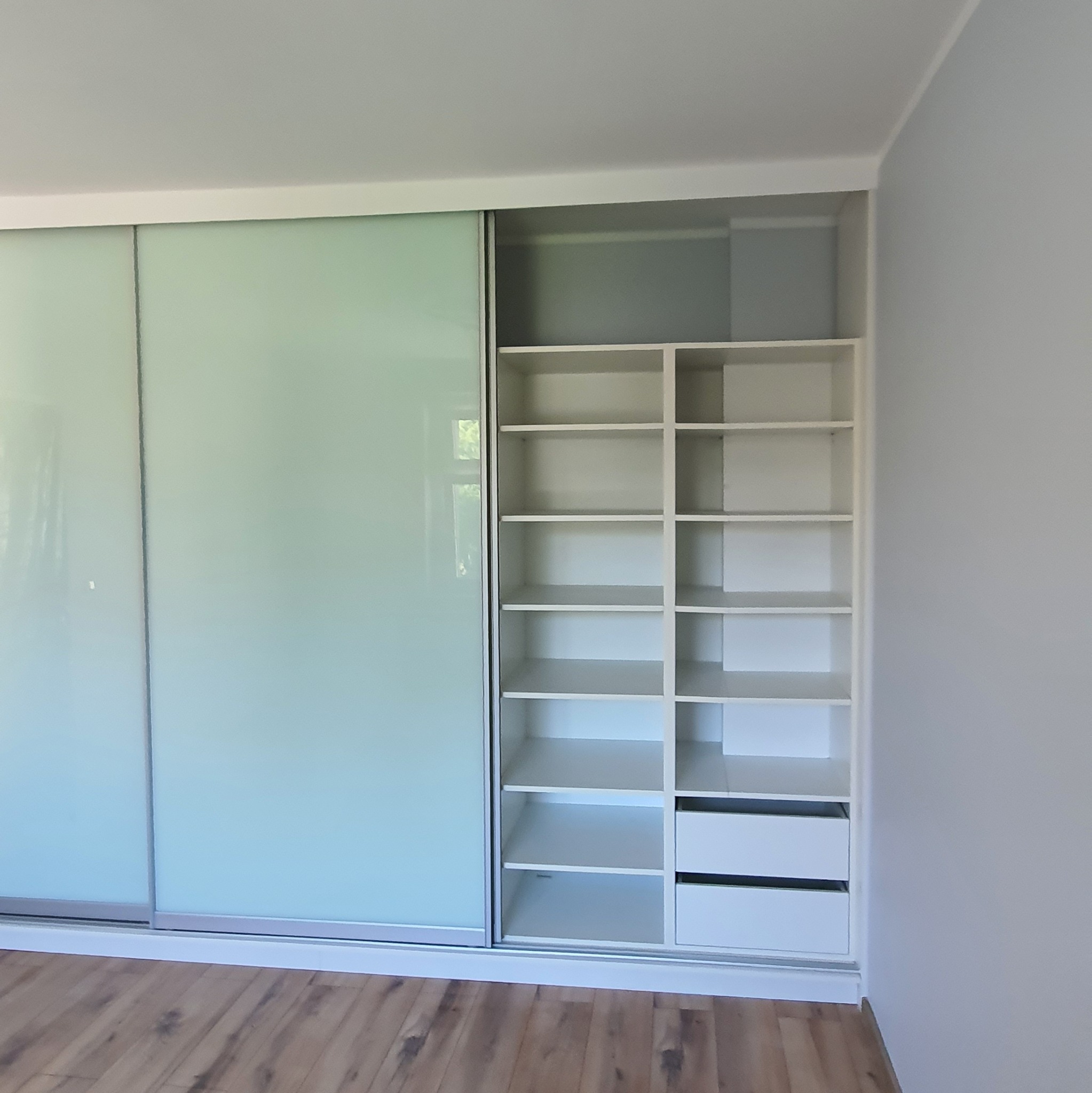 Nowoczesna, biała szafa wnękowa z przesuwnymi drzwiami z mlecznego szkła, wkomponowana w jasne wnętrze. Widoczne półki i szuflady.