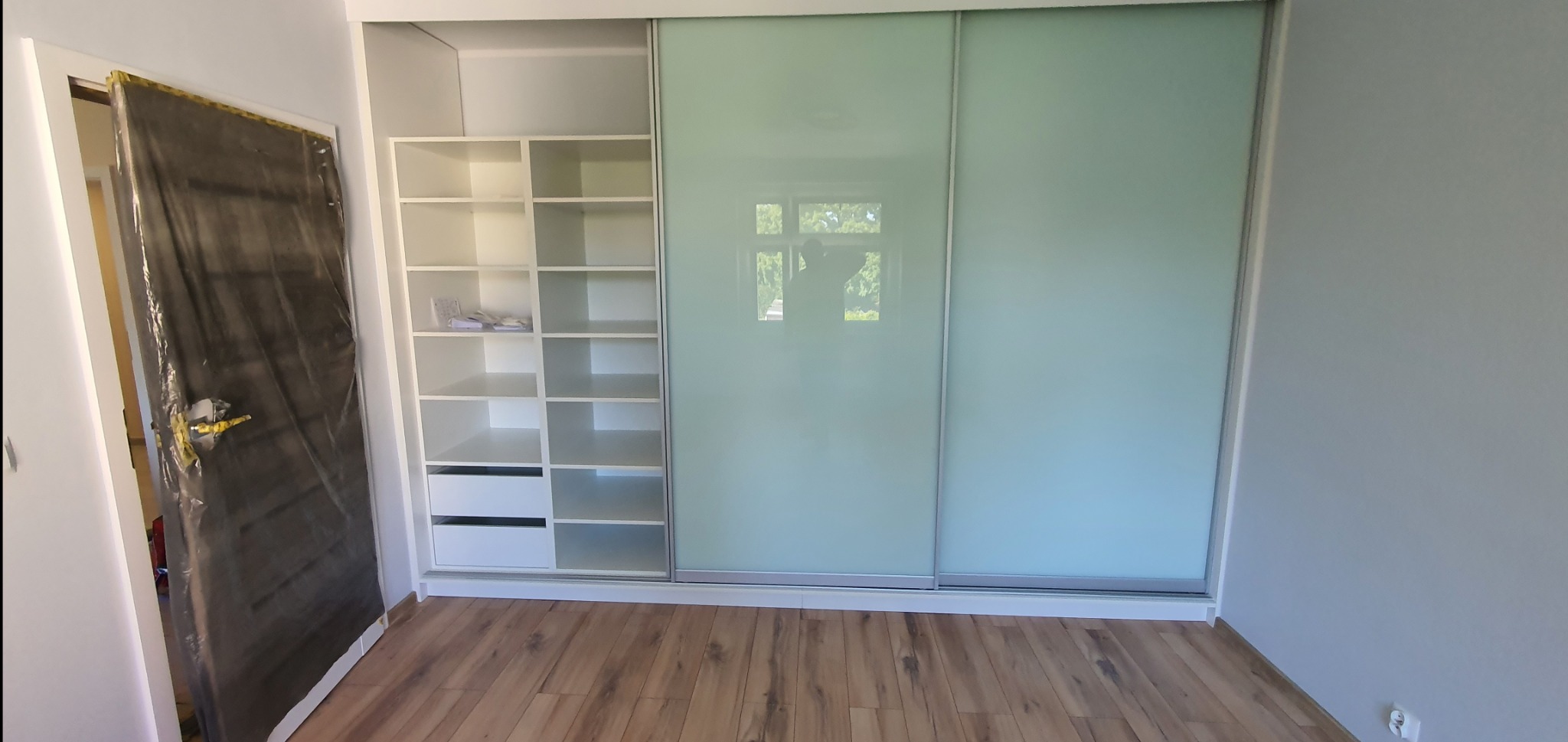 Wnętrze z zabudową: biały regał z półkami i szufladami oraz szafa przesuwna z mlecznymi szybami. Podłoga z paneli drewnopodobnych. Drzwi zabezpieczone folią.