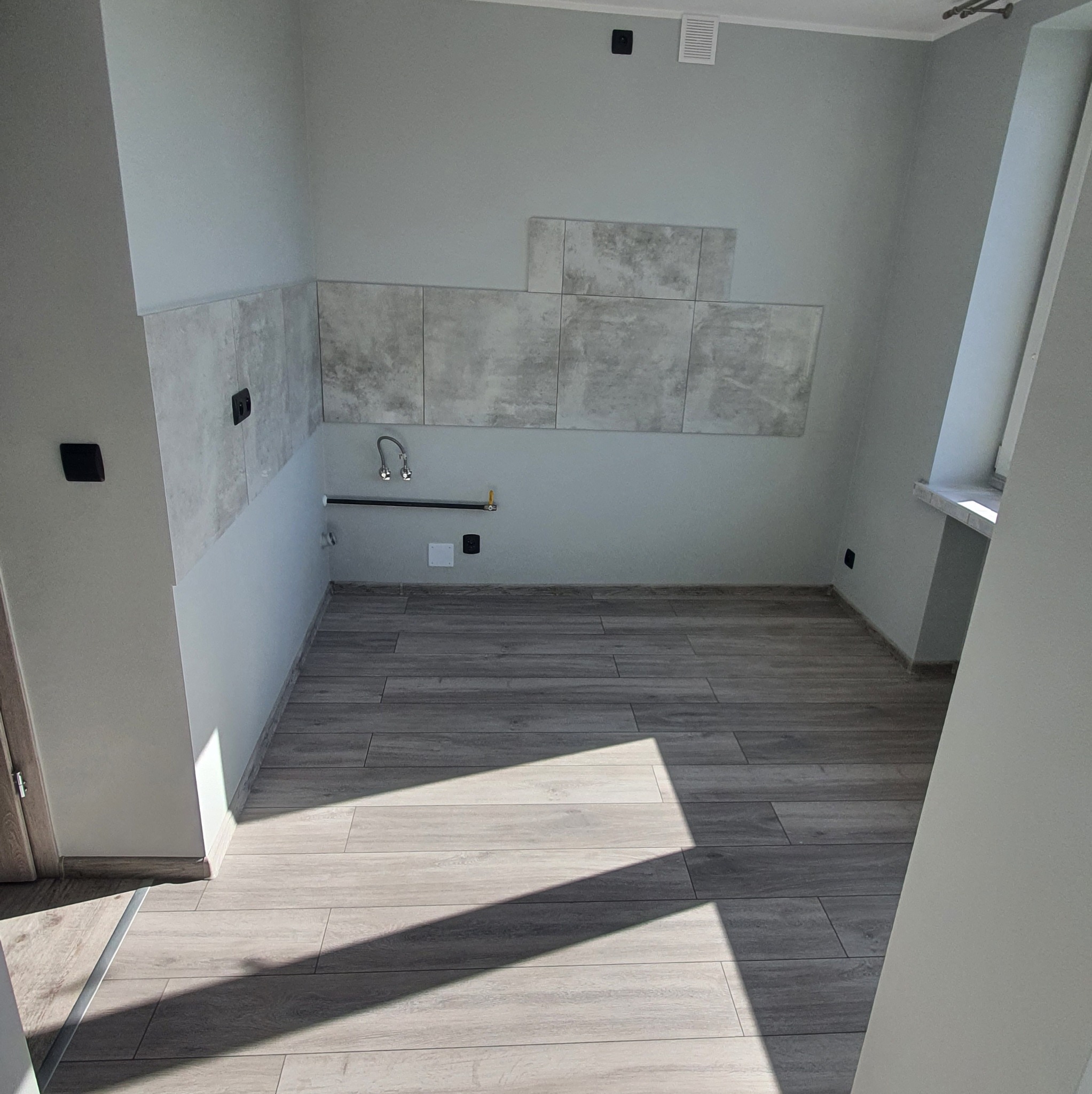 Jasna łazienka w trakcie wykańczania: położona podłoga z płytek imitujących drewno, częściowo ułożone płytki na ścianie, widoczne instalacje wodne i okno.