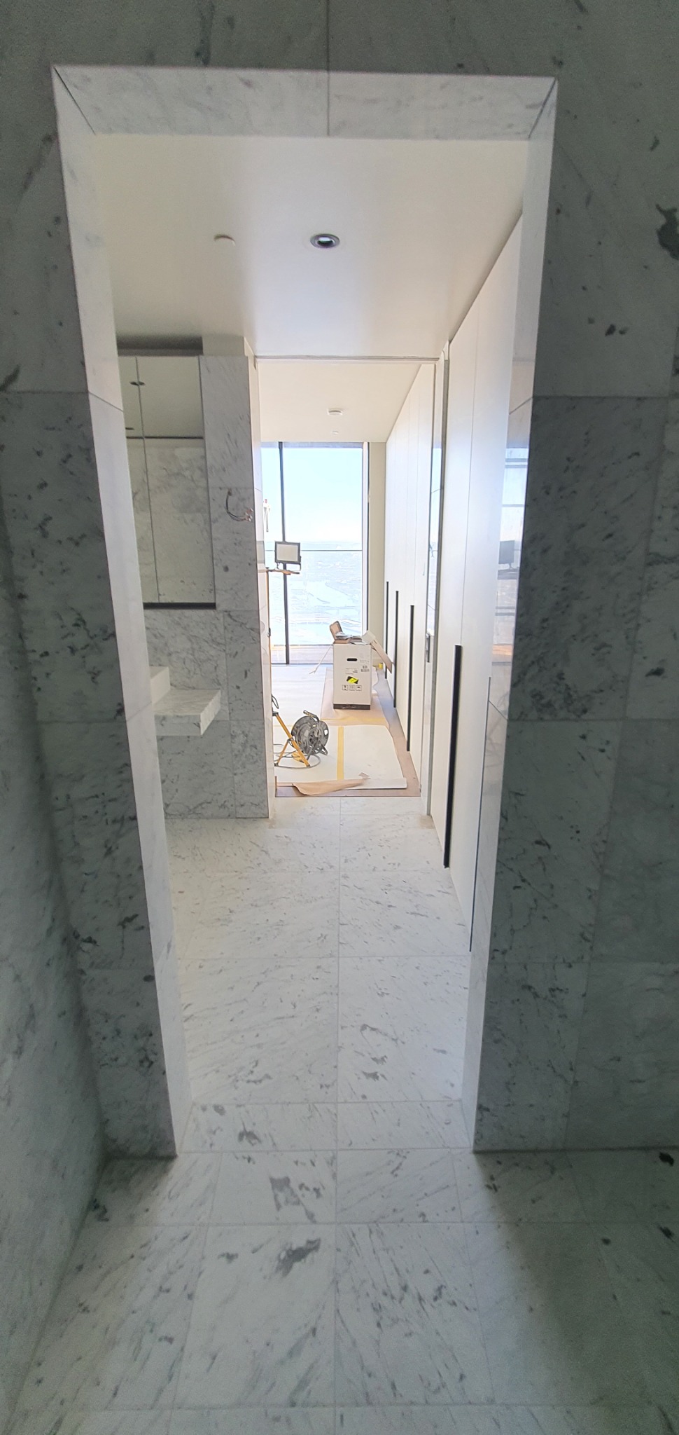 Wykończenie łazienki marmurem: widok przez otwór w ścianie na posadzkę i fragment szafy. W tle narzędzia i widok na miasto. Nowoczesny design, jasne wnętrze.