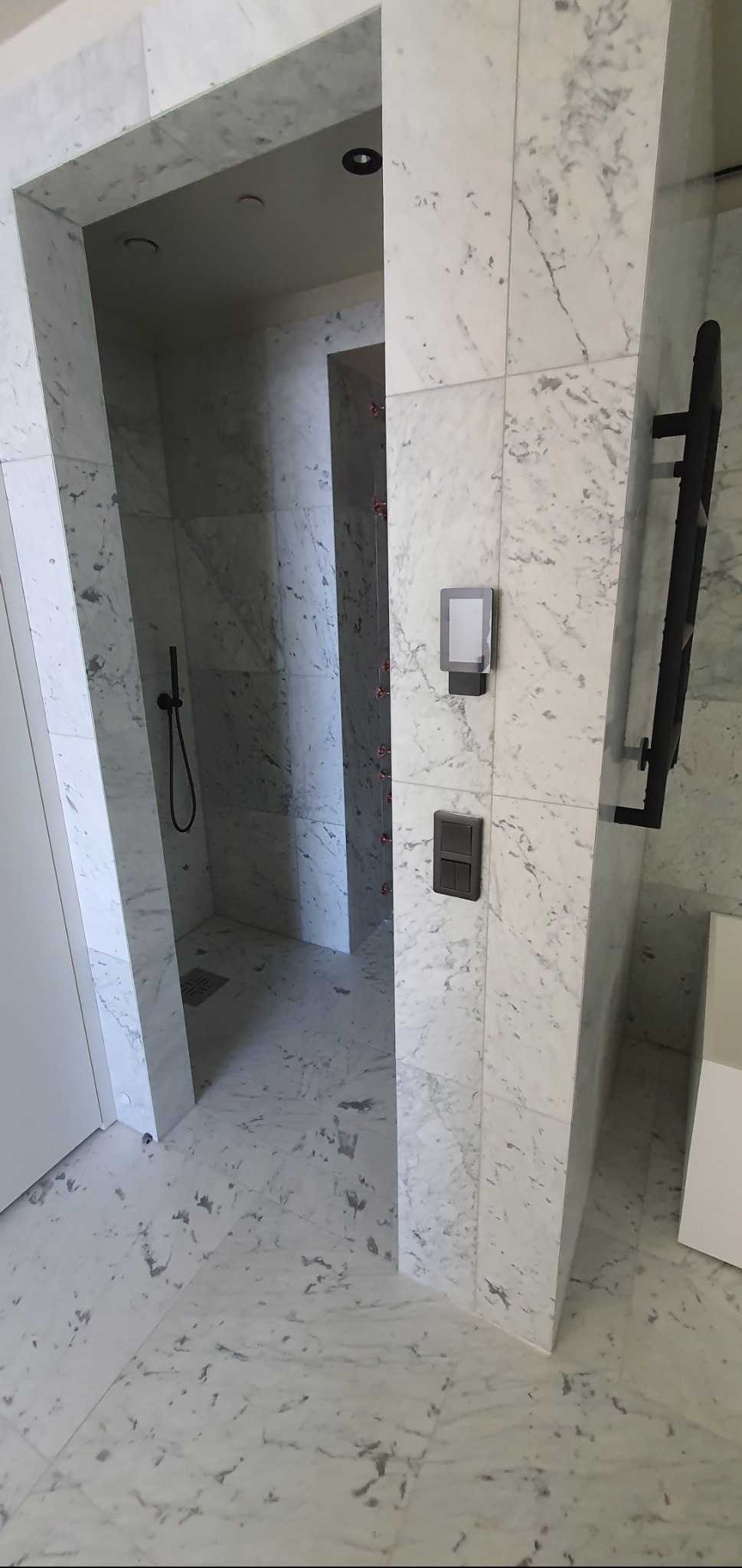 Nowoczesna łazienka z marmurowym wykończeniem ścian i podłogi. Widoczny prysznic z czarną armaturą, oświetlenie i grzejnik.