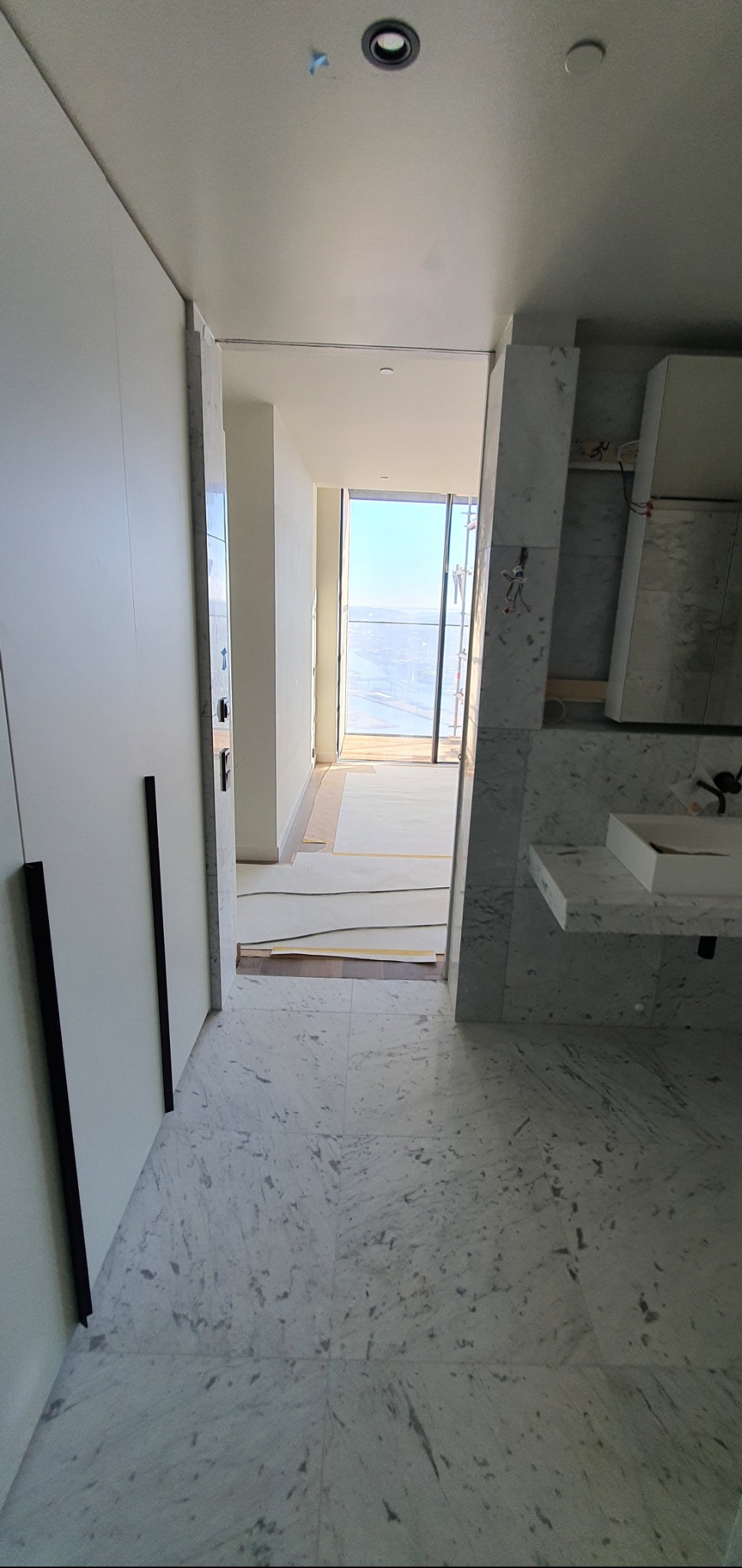 Wnętrze łazienki w trakcie wykończenia: białe marmurowe płytki podłogowe, zabudowa meblowa z czarnymi uchwytami i widok na balkon zza osłoniętej podłogi.