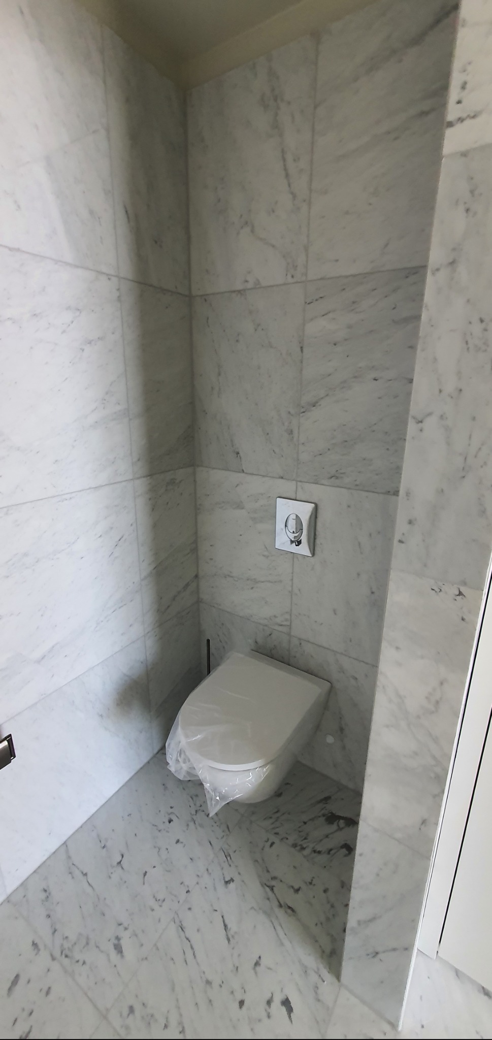 Wykończona łazienka z marmurowymi płytkami, wiszącą toaletą zabezpieczoną folią i minimalistycznym przyciskiem spłukującym na ścianie.