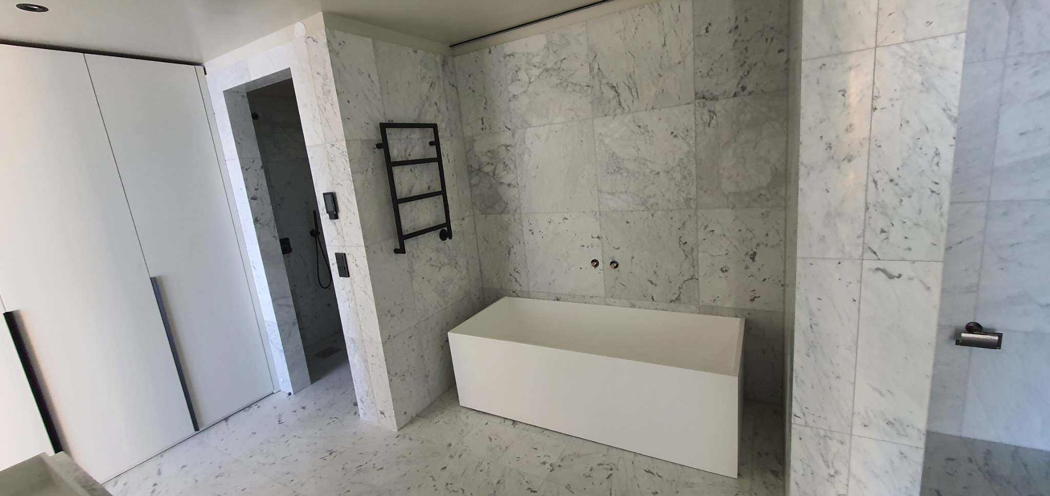 Nowoczesna łazienka wykończona białym marmurem z minimalistyczną wanną i czarnym grzejnikiem drabinkowym. Widoczna kabina prysznicowa.