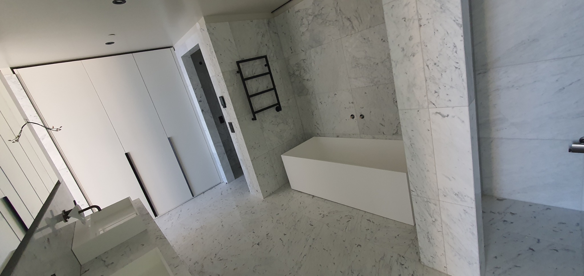 Nowoczesna łazienka z marmurowymi płytkami, białą wanną i szafkami. Minimalistyczny design z akcentami czerni. Widok na umywalkę i grzejnik drabinkowy.