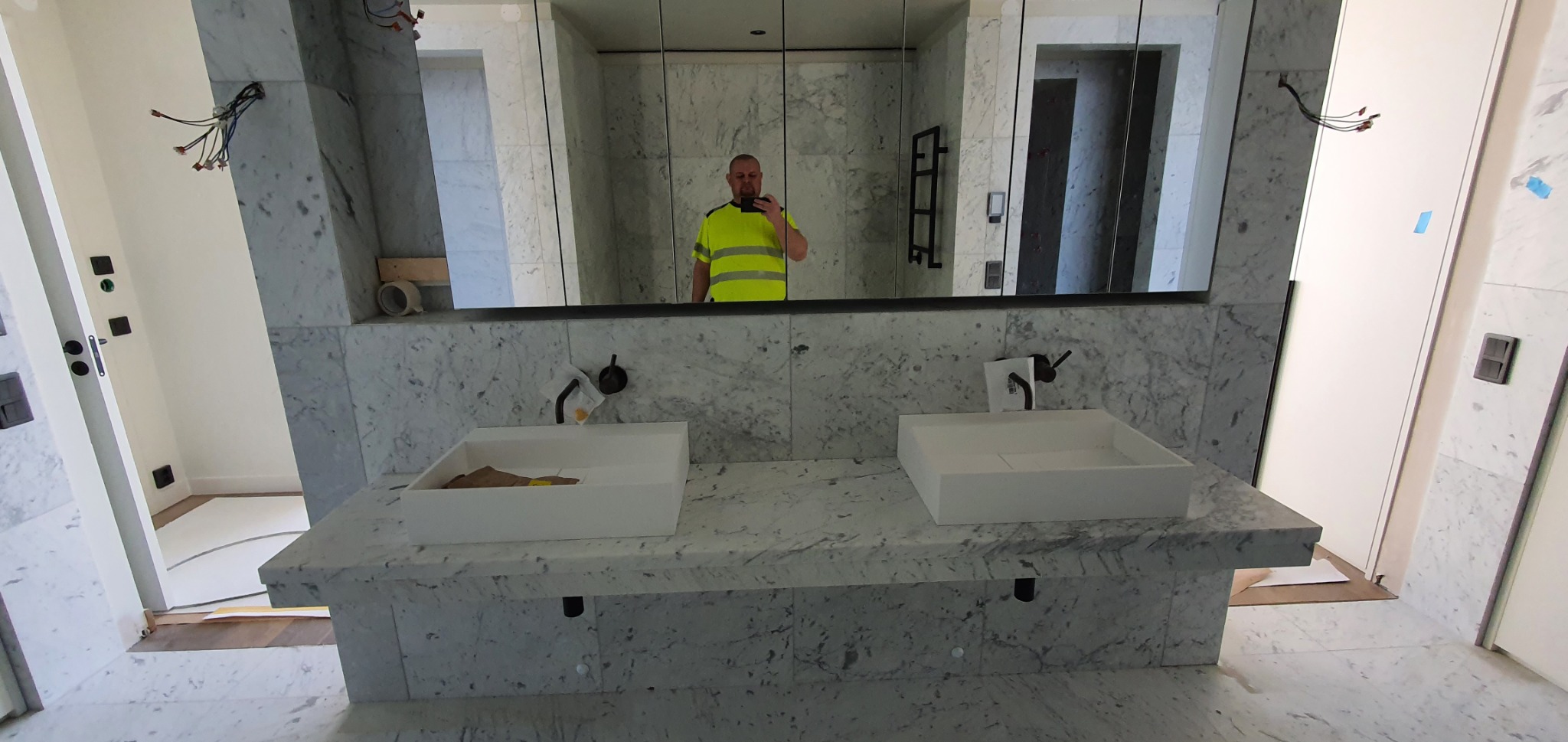 Łazienka w trakcie wykończenia z umywalkami z białego kamienia na marmurowym blacie. W lustrze odbija się osoba w żółtej kamizelce.