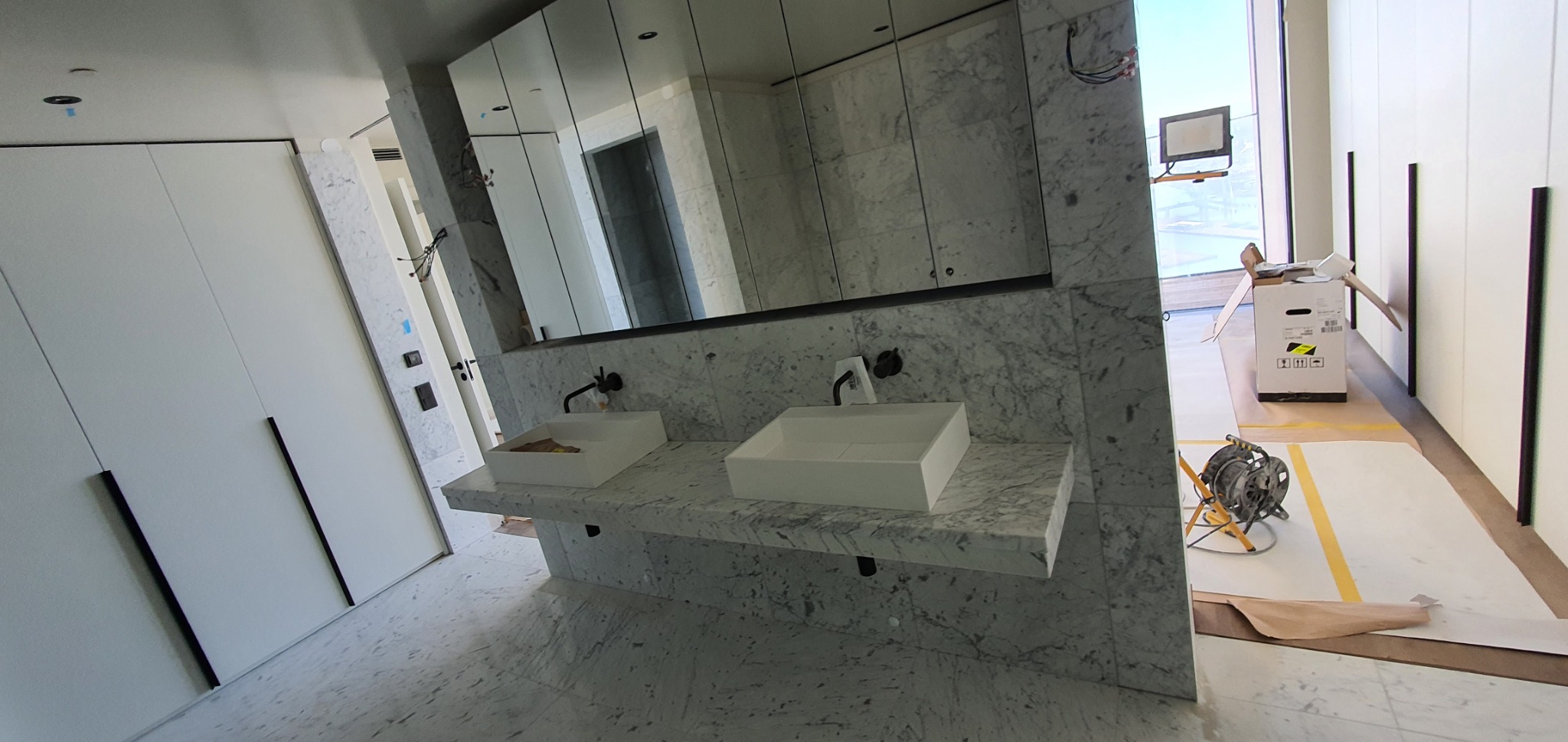Nowoczesna łazienka z marmurową ścianą i podwójną umywalką. Widoczne duże lustro i białe szafy. Na podłodze zabezpieczenia i sprzęt budowlany.