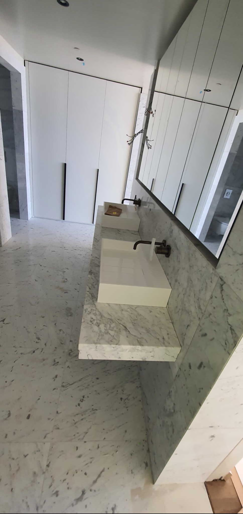 Nowoczesna łazienka z marmurową posadzką i blatem, minimalistyczna armatura w kolorze brązu, białe szafki w zabudowie i duże lustro.