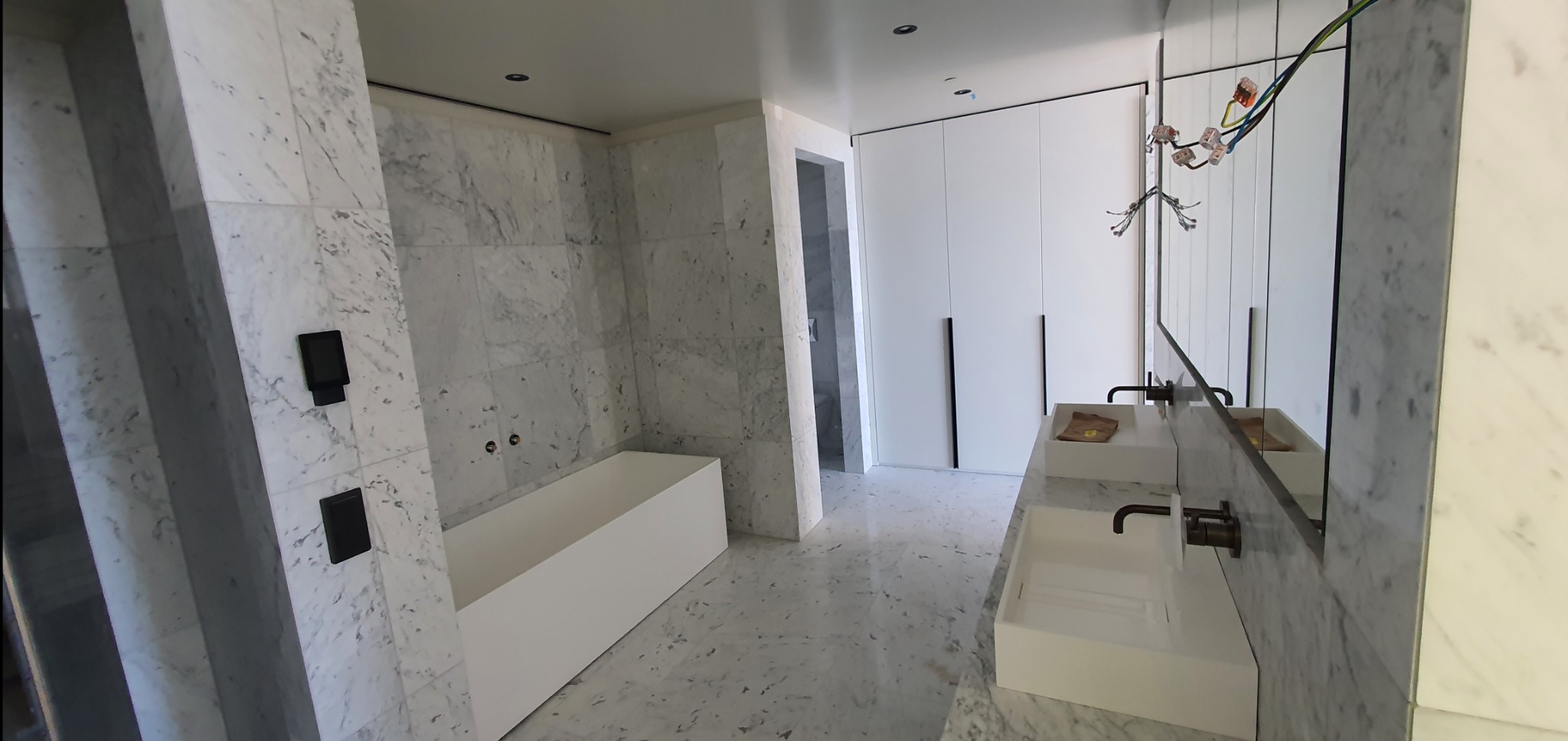 Elegancka, minimalistyczna łazienka z marmurowymi ścianami i podłogą. Biała wanna i umywalki o prostych kształtach. Widoczne instalacje elektryczne przy lustrze.