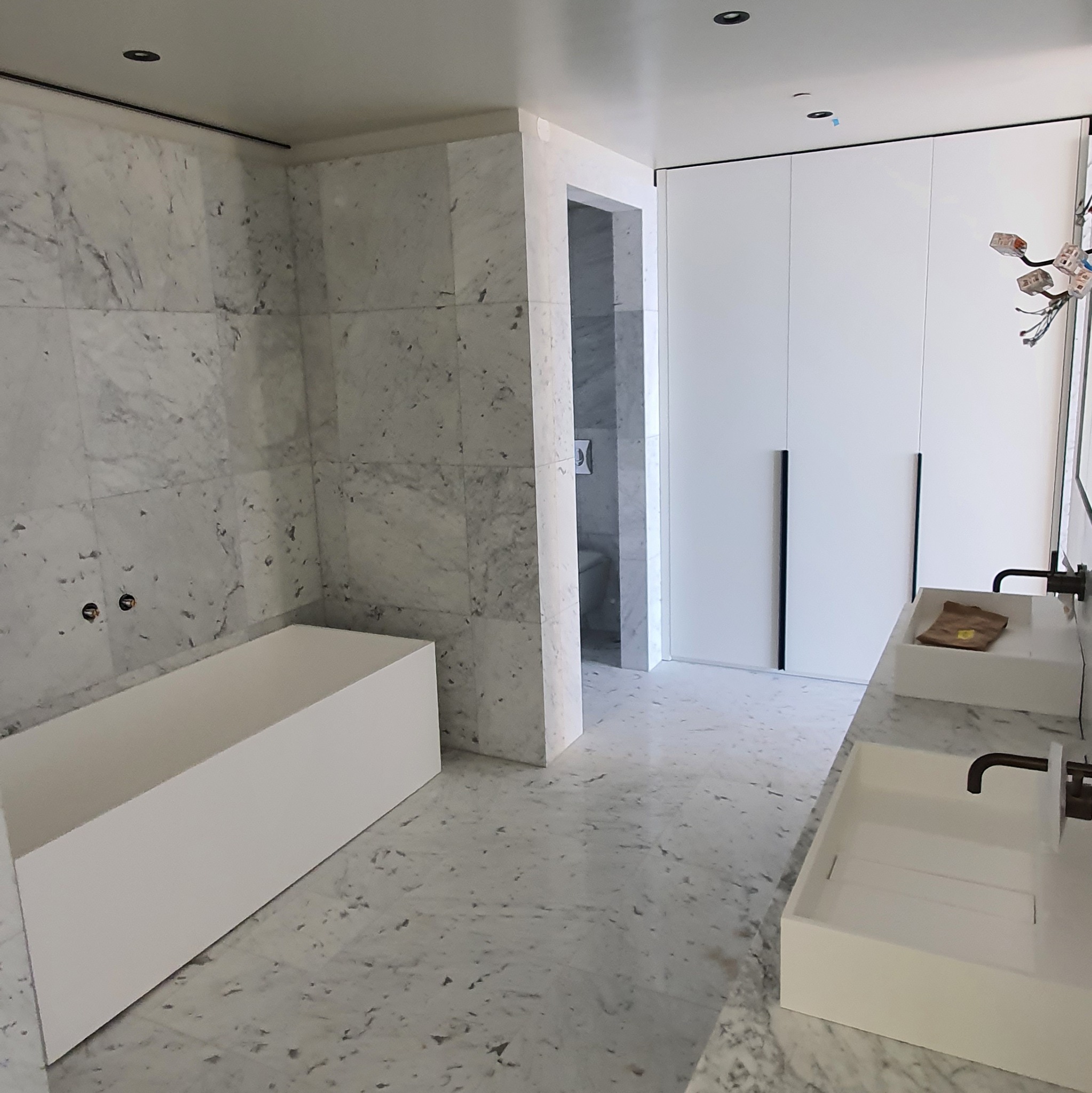 Nowoczesna łazienka z marmurowymi płytkami, białą wanną i umywalkami. Widoczne elementy instalacji elektrycznej. Minimalistyczny design, jasne wnętrze.