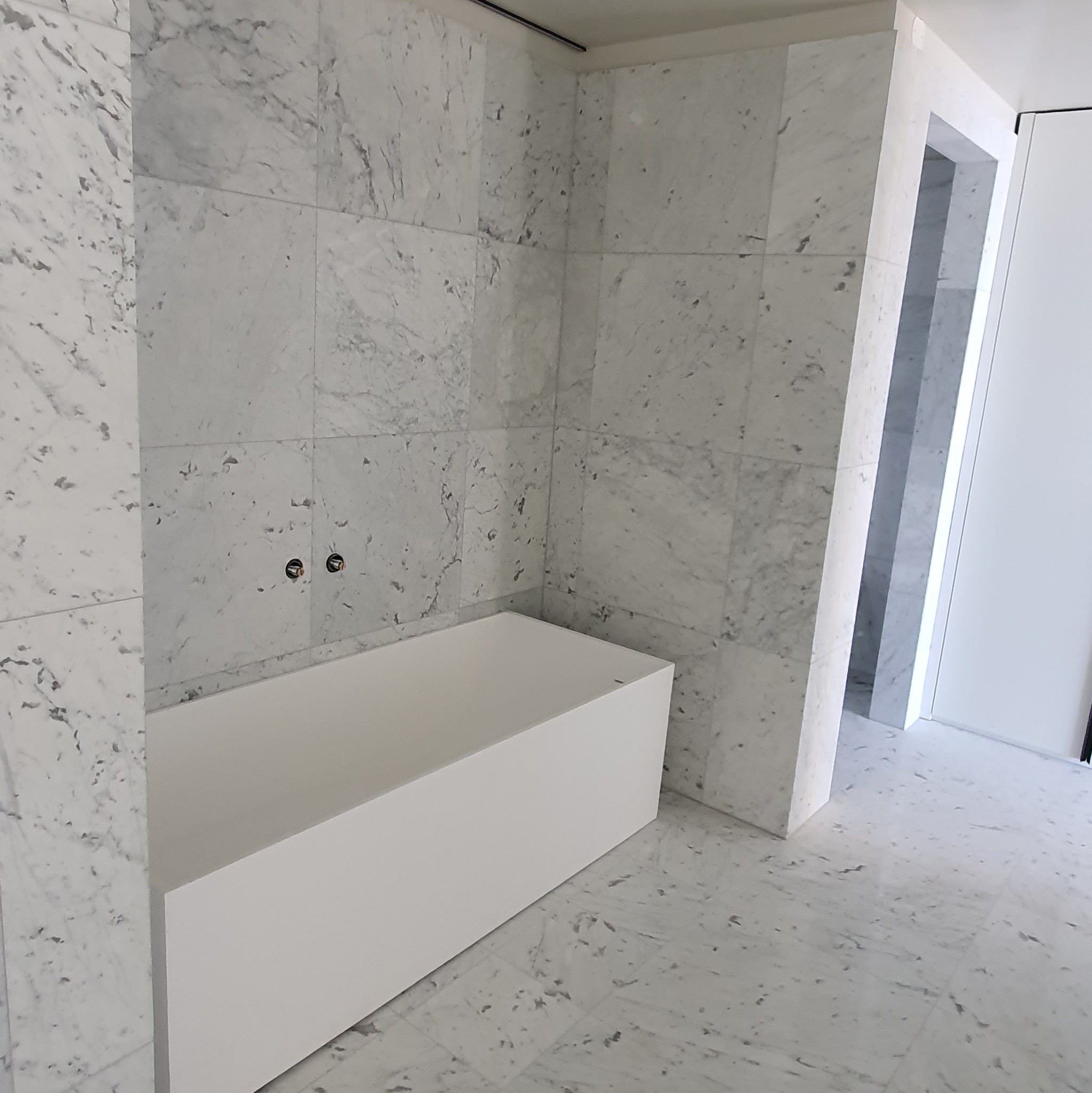 Minimalistyczna łazienka z wanną obłożona białym marmurem z szarymi żyłami, widoczne przyłącza baterii. Eleganckie wykończenie w jasnych barwach.