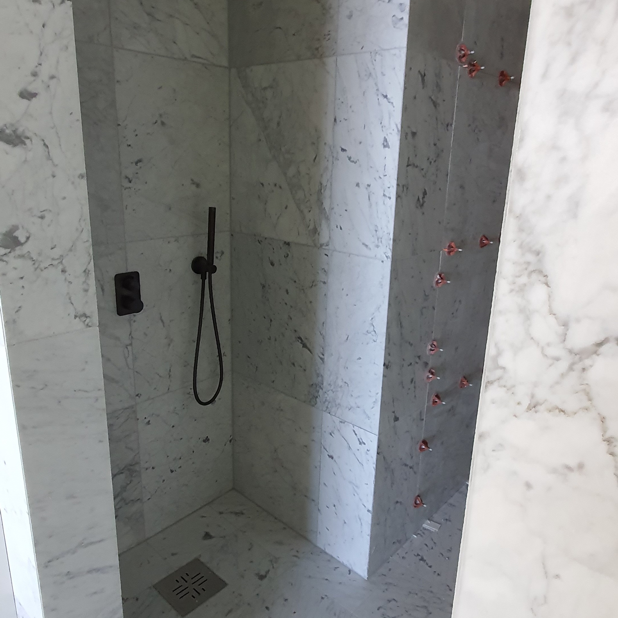 Wykończona kabina prysznicowa z marmurowymi płytkami, czarną armaturą i odpływem liniowym. Na ścianie widoczne krzyżyki dystansowe do płytek.