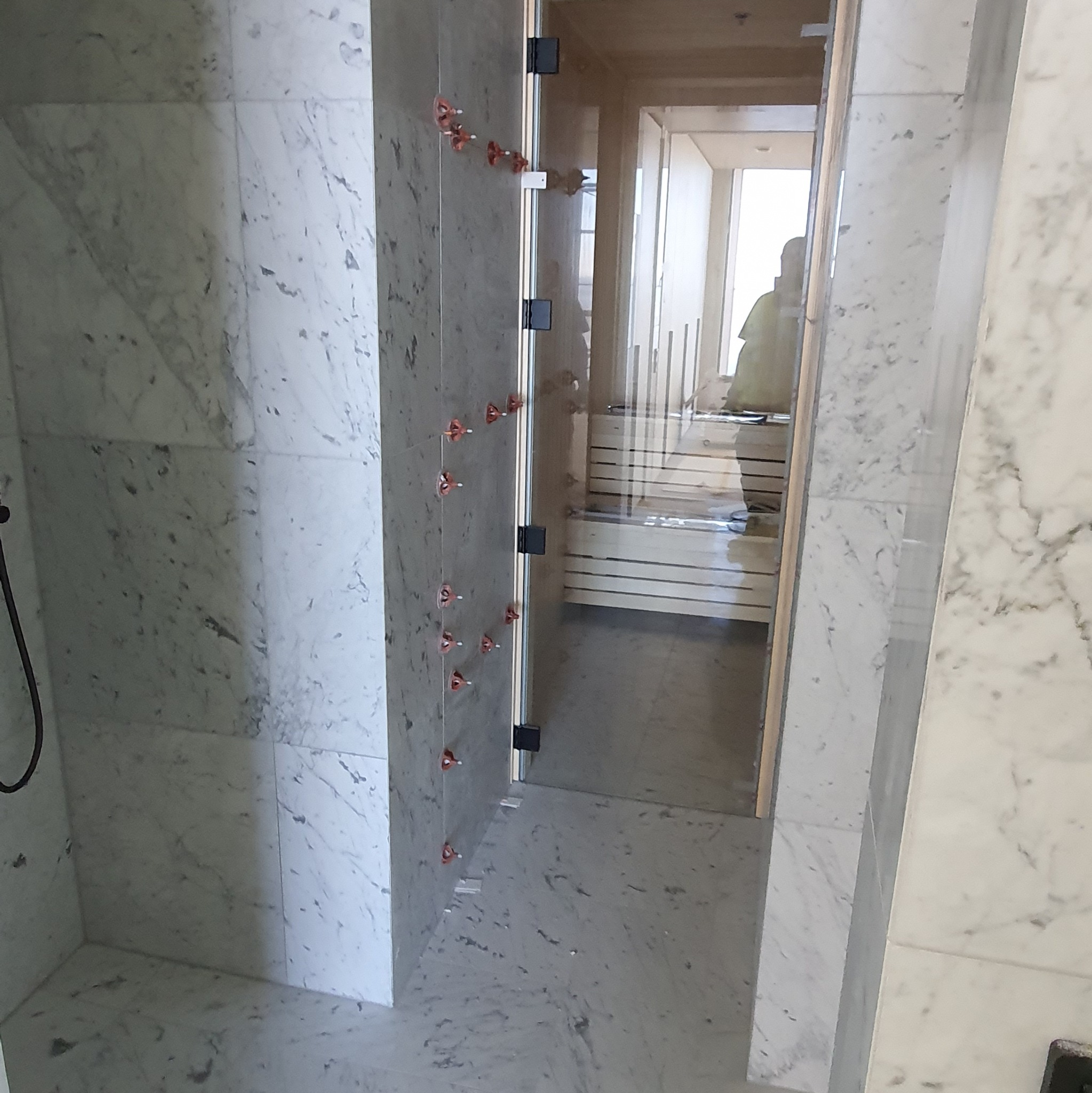 Biała marmurowa łazienka z kabiną prysznicową w trakcie montażu, widoczne krzyżyki glazurnicze. W tle widoczna sauna.