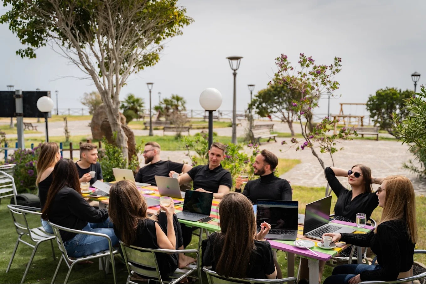 Zespół w czarnych ubraniach pracuje przy kolorowym stole na zewnątrz, używając laptopów, pijąc napoje. W tle park i morze. Współpraca w plenerze.